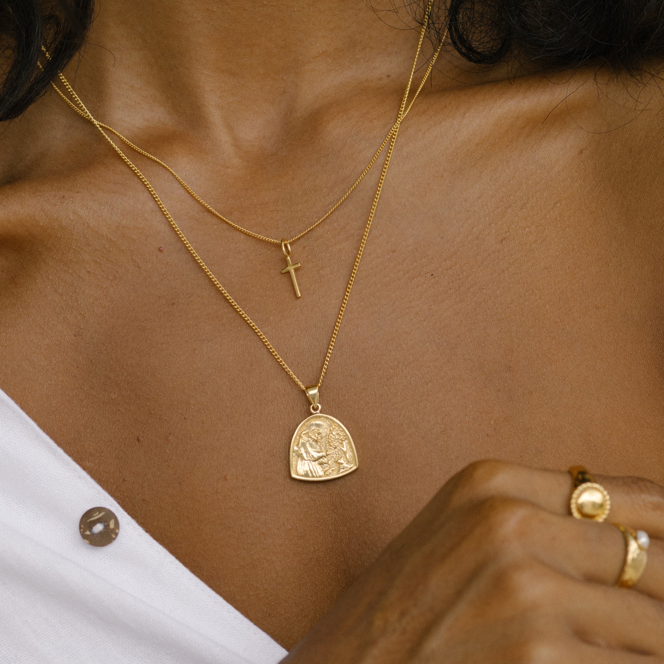 A close-up showing two layered gold necklaces: one with a small cross pendant and another with a larger medallion, worn with a white shirt.