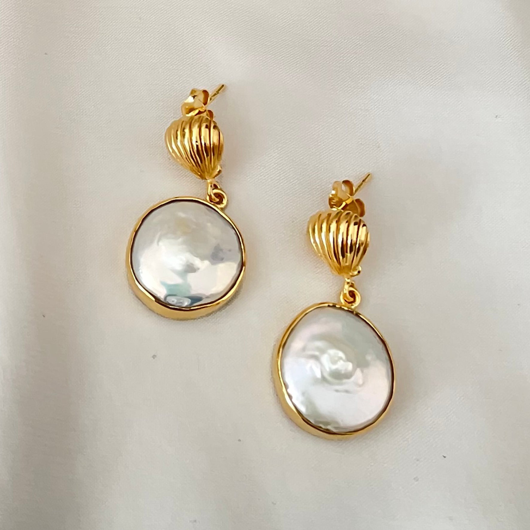 A pair of gold drop earrings with large, round white pearl pendants displayed on white fabric.
