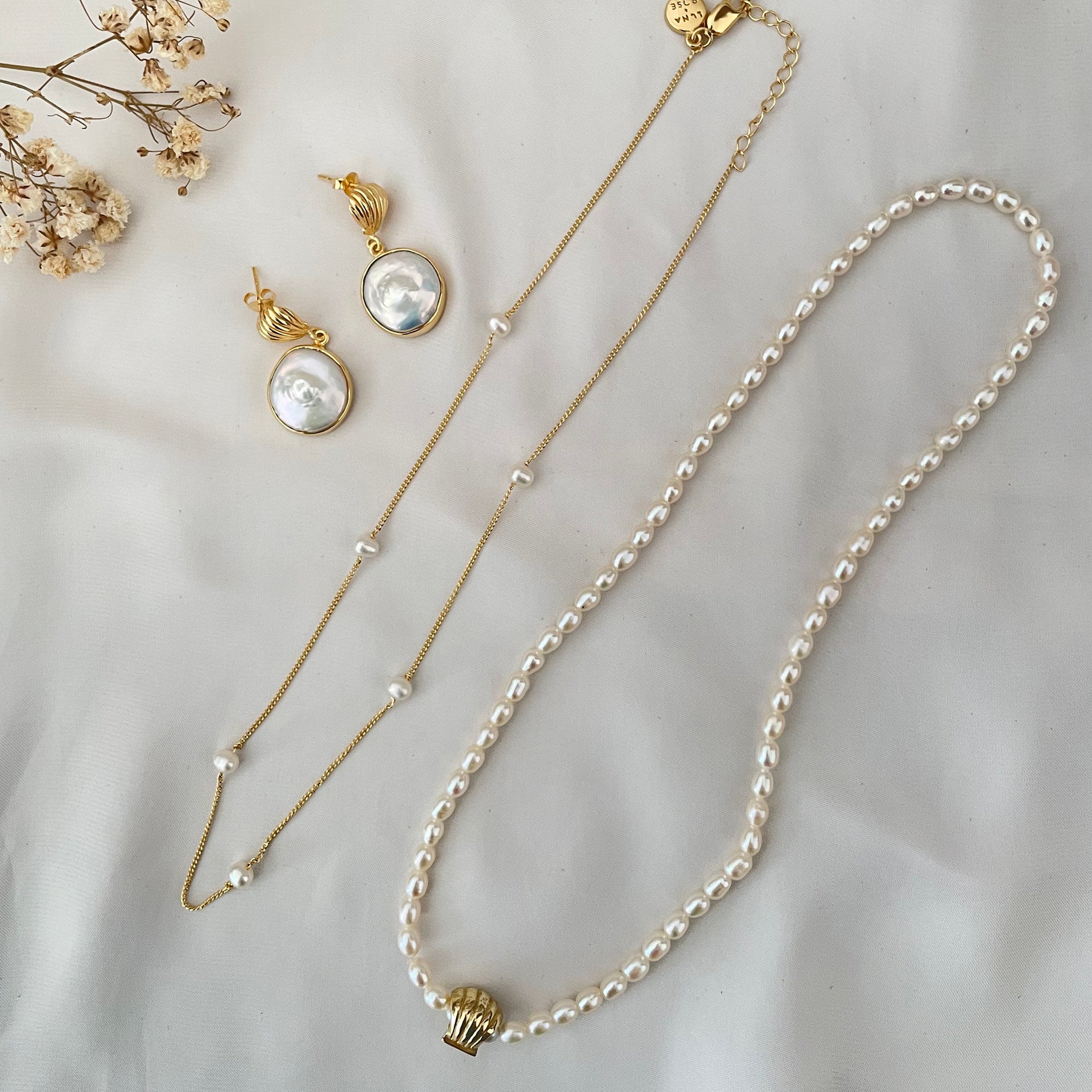 Gold and pearl jewelry pieces, including a pair of drop earrings, a gold necklace with pearl accents, and a pearl necklace with a gold clasp, arranged on a white fabric