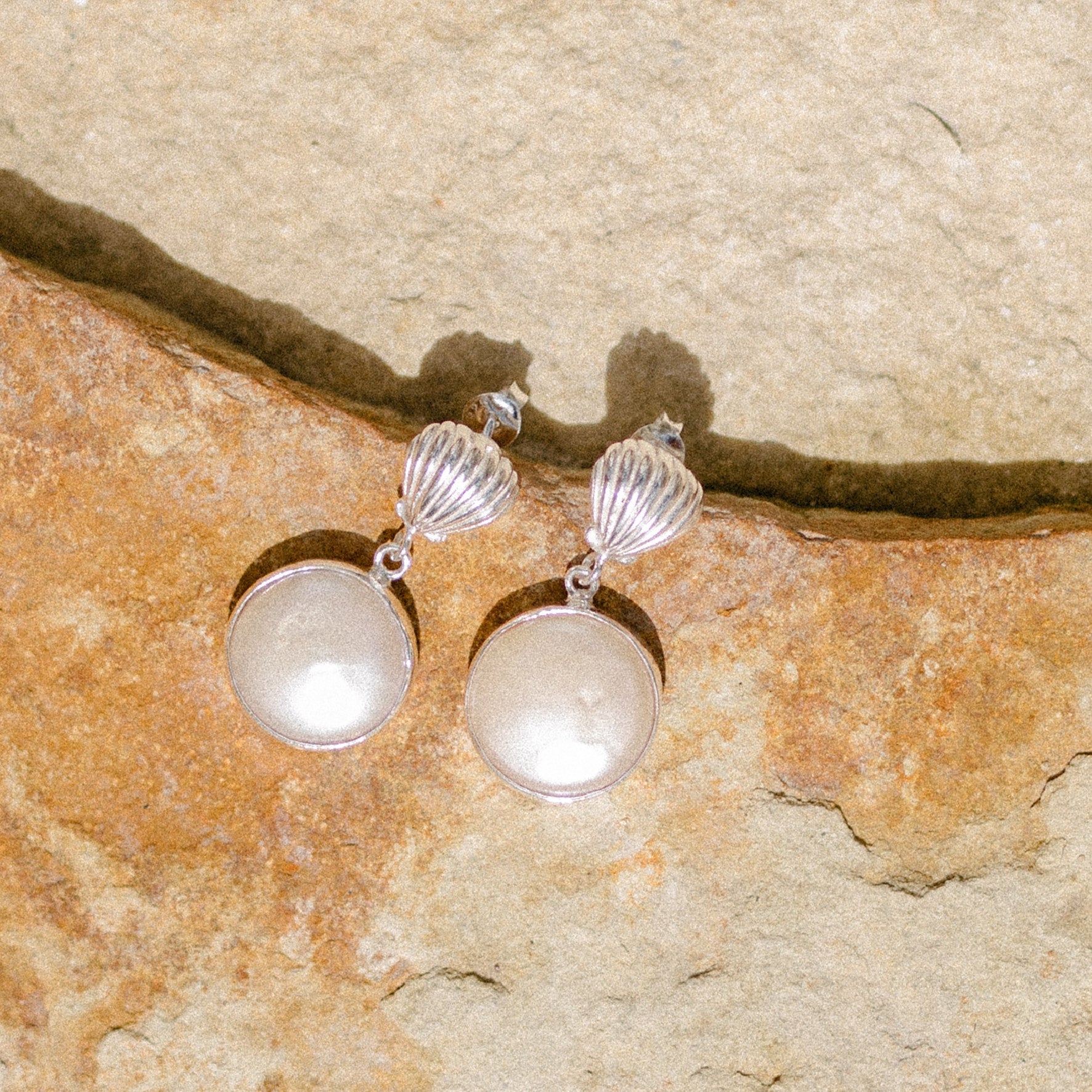 A pair of silver shell earrings with large, round white pearl pendants displayed on a stone