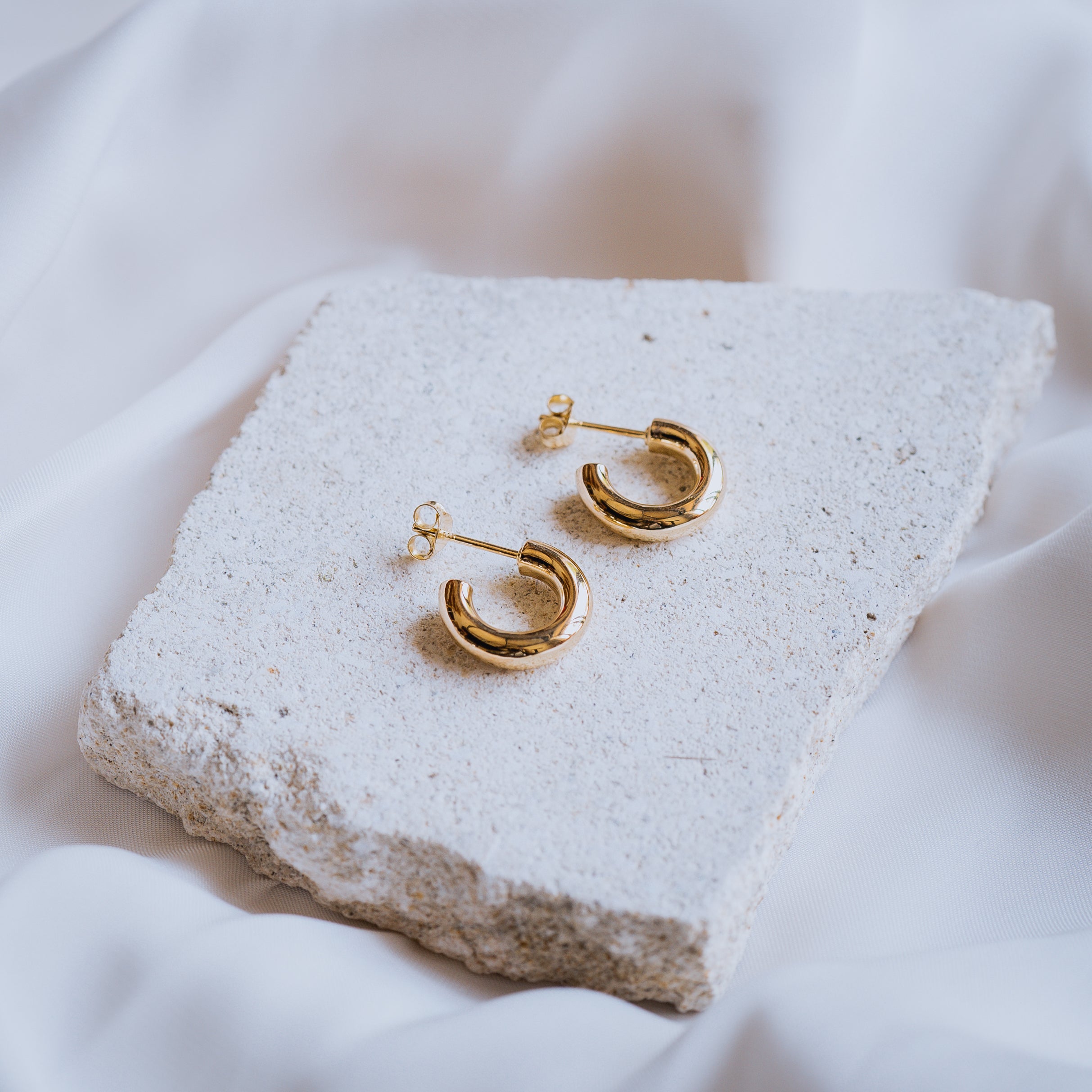 A pair of gold hoop earrings with post backs displayed on a textured beige stone
