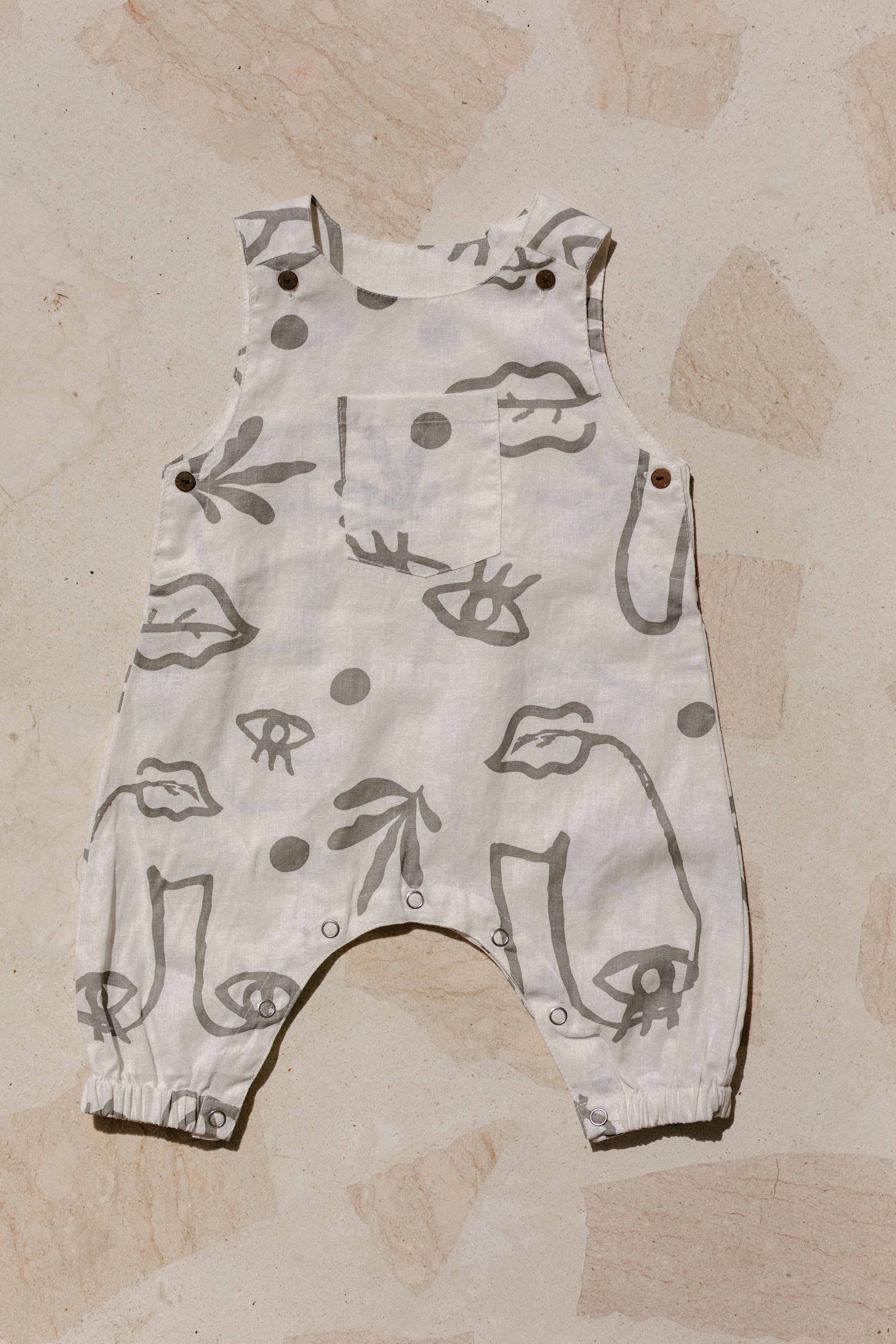 A white/grey romper with grey abstract face designs and snap buttons along the legs and shoulders is laid flat.
