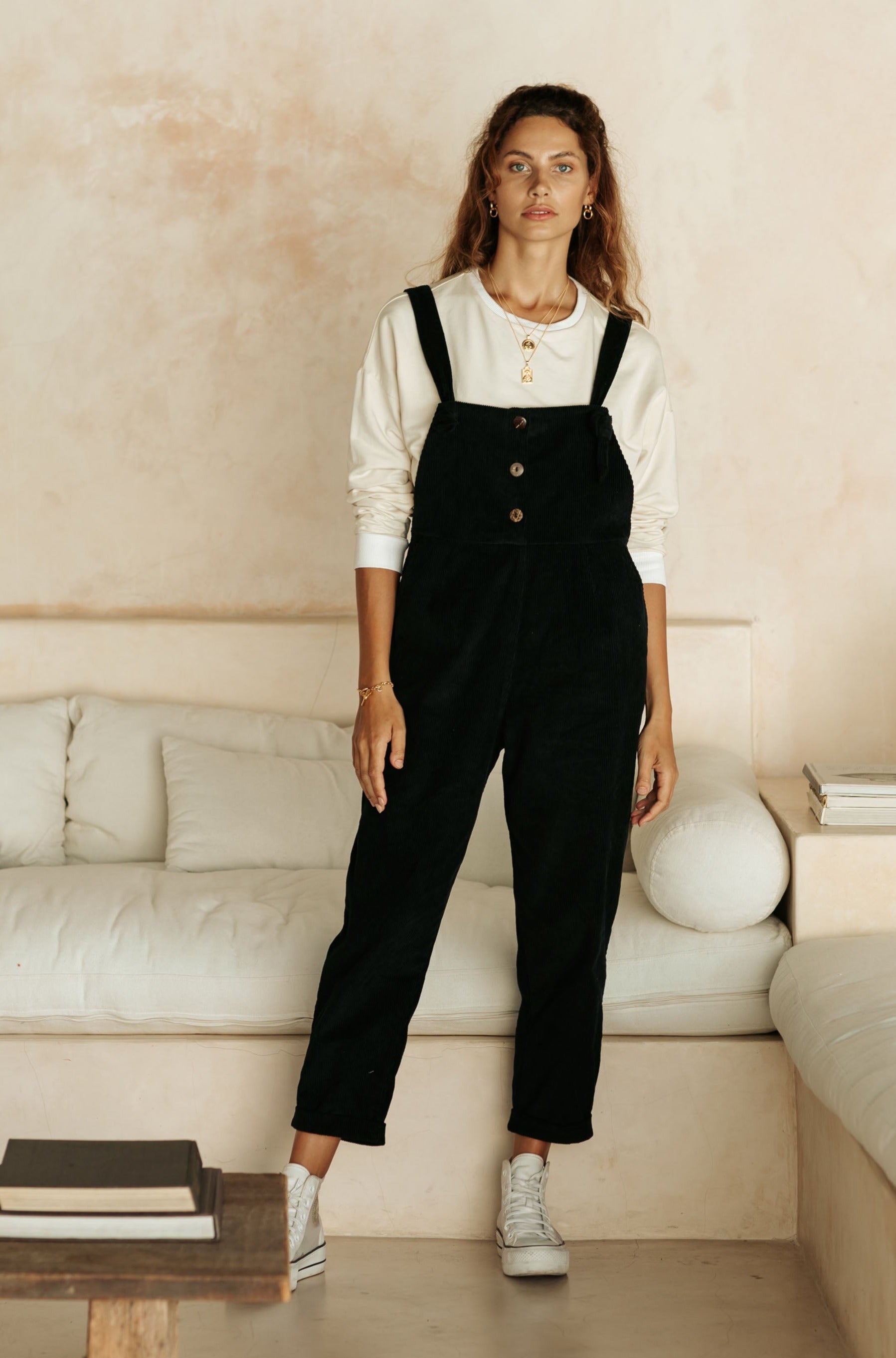 A woman in black overalls and a white top, wearing white sneakers, stands in front of a white couch and a minimalist background.