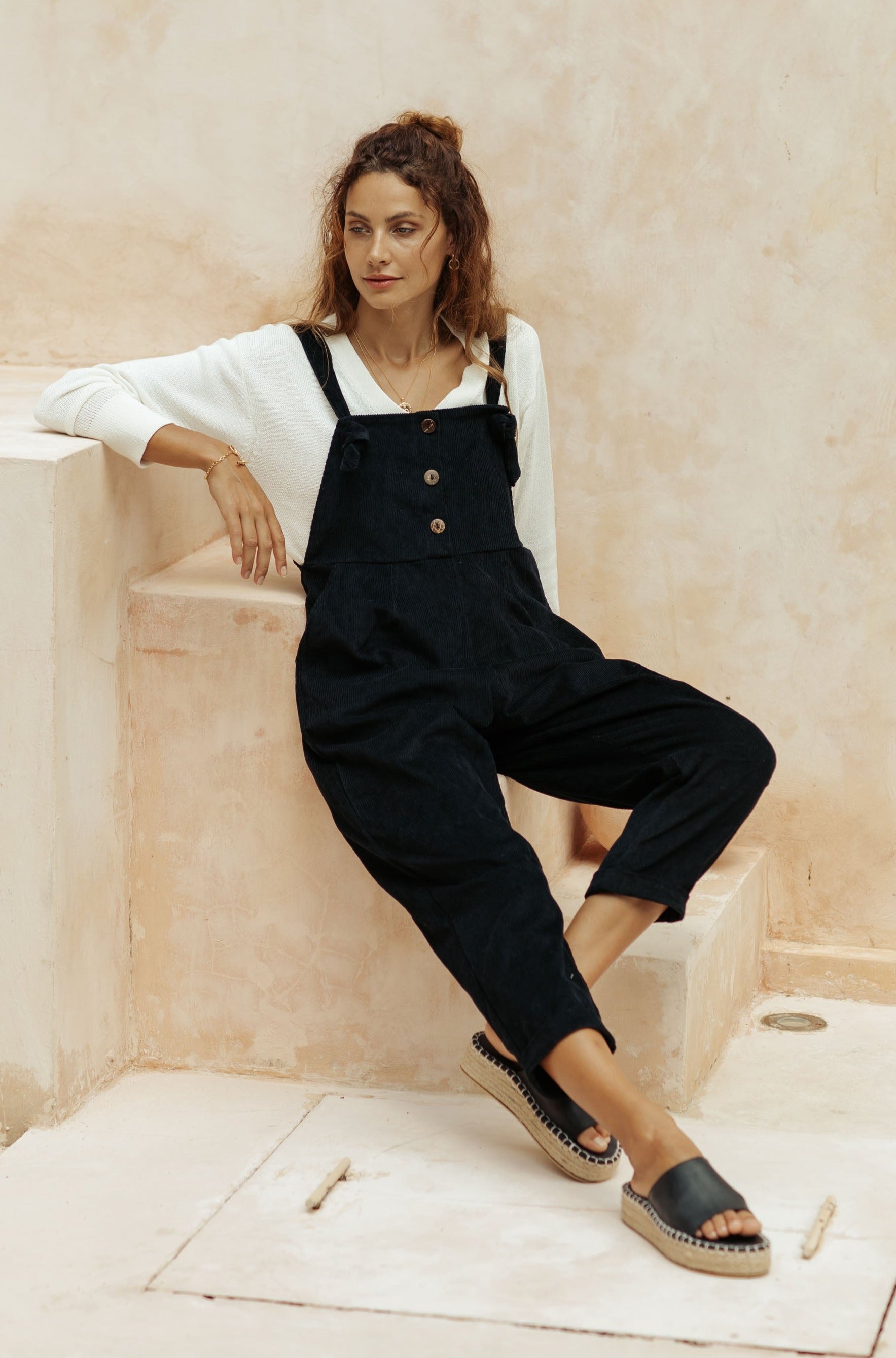 A woman dressed in black overalls with a white top, sitting on stairs in a minimalistic space.
