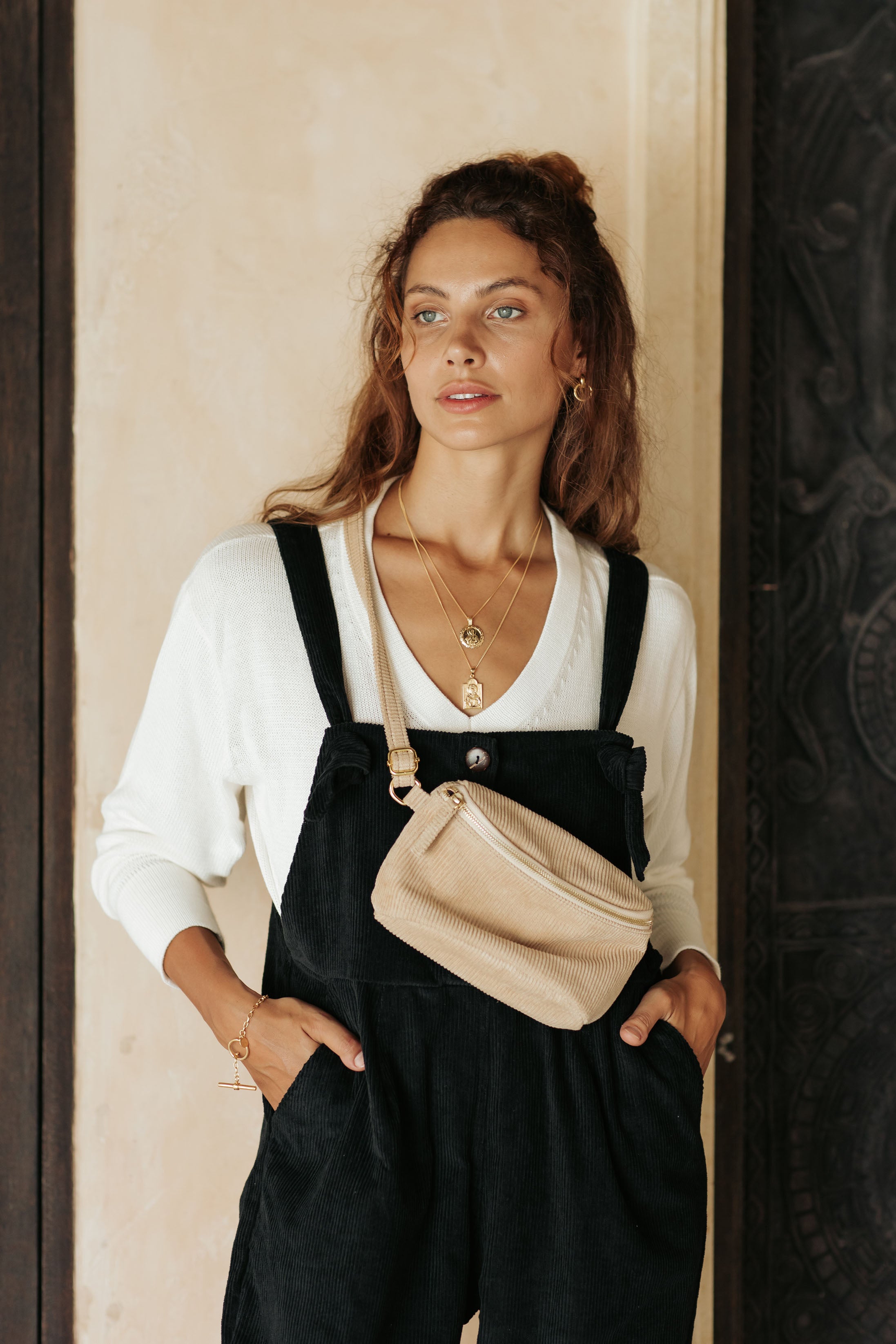 A woman wearing black overalls and a white top, accessorized with a beige fanny pack, stands with her hands in her pockets.