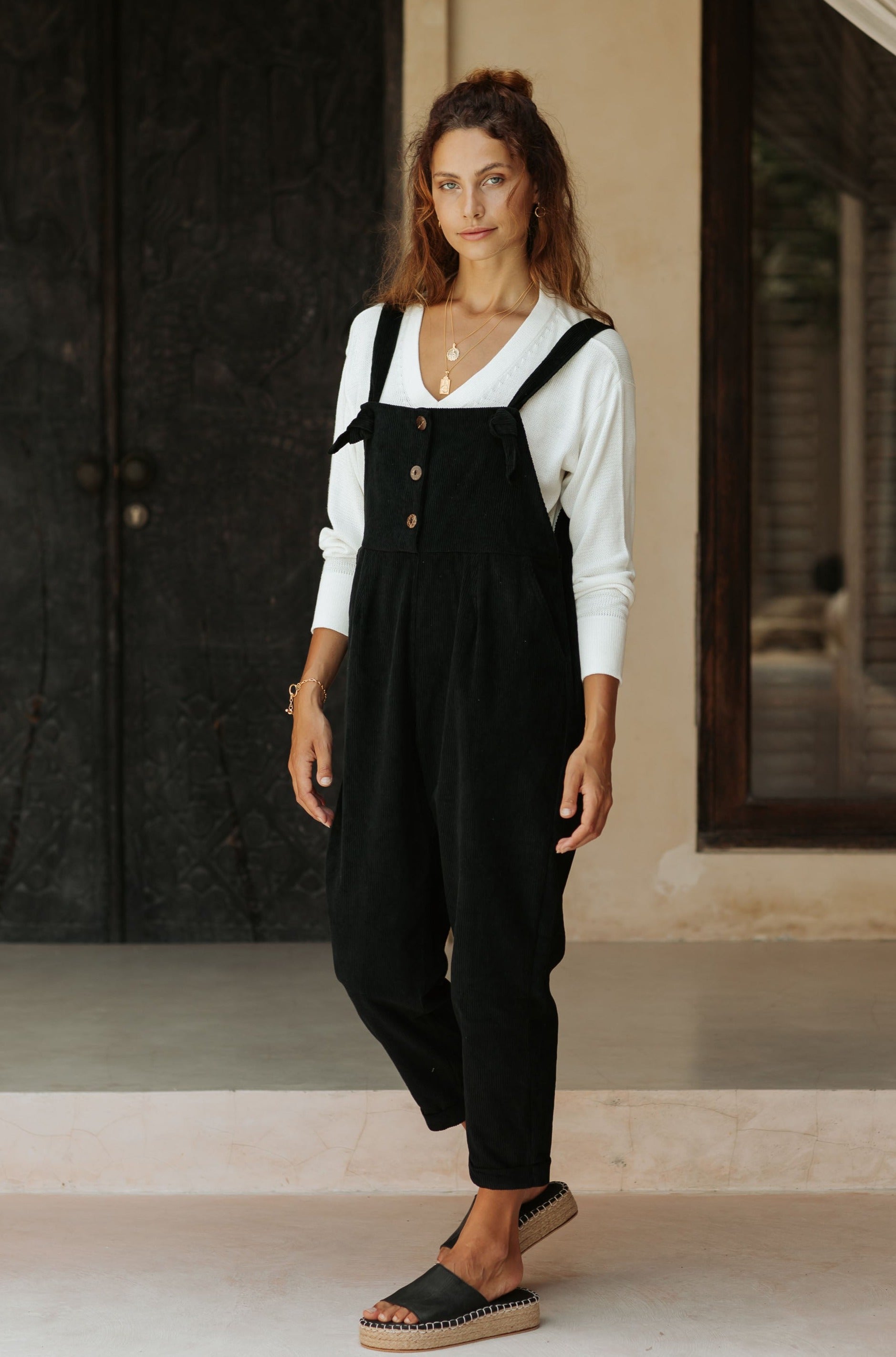 A woman in black overalls and a white top stands in a room with a neutral color palette and textured walls.