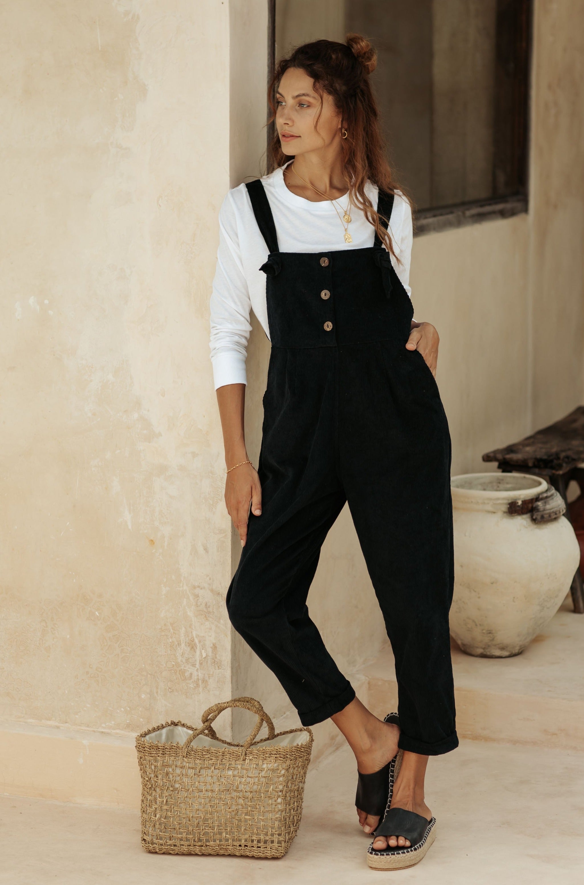 A woman in black overalls and a white top leans against a wall, next to a wicker basket in a neutral-toned setting.