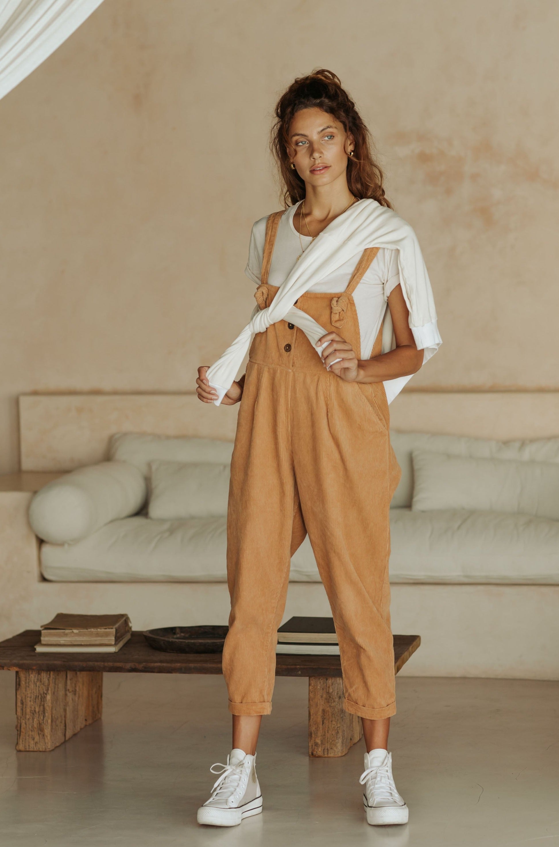 A woman in tan overalls and a white t-shirt with a sweater draped over her shoulders, standing in a room with neutral decor.