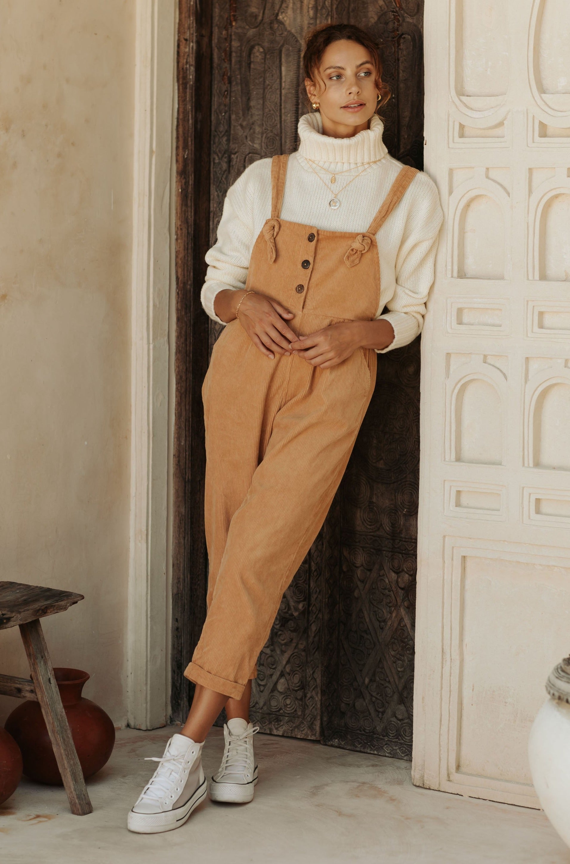 A woman in tan overalls and a white turtleneck sweater, standing against a rustic wooden door.