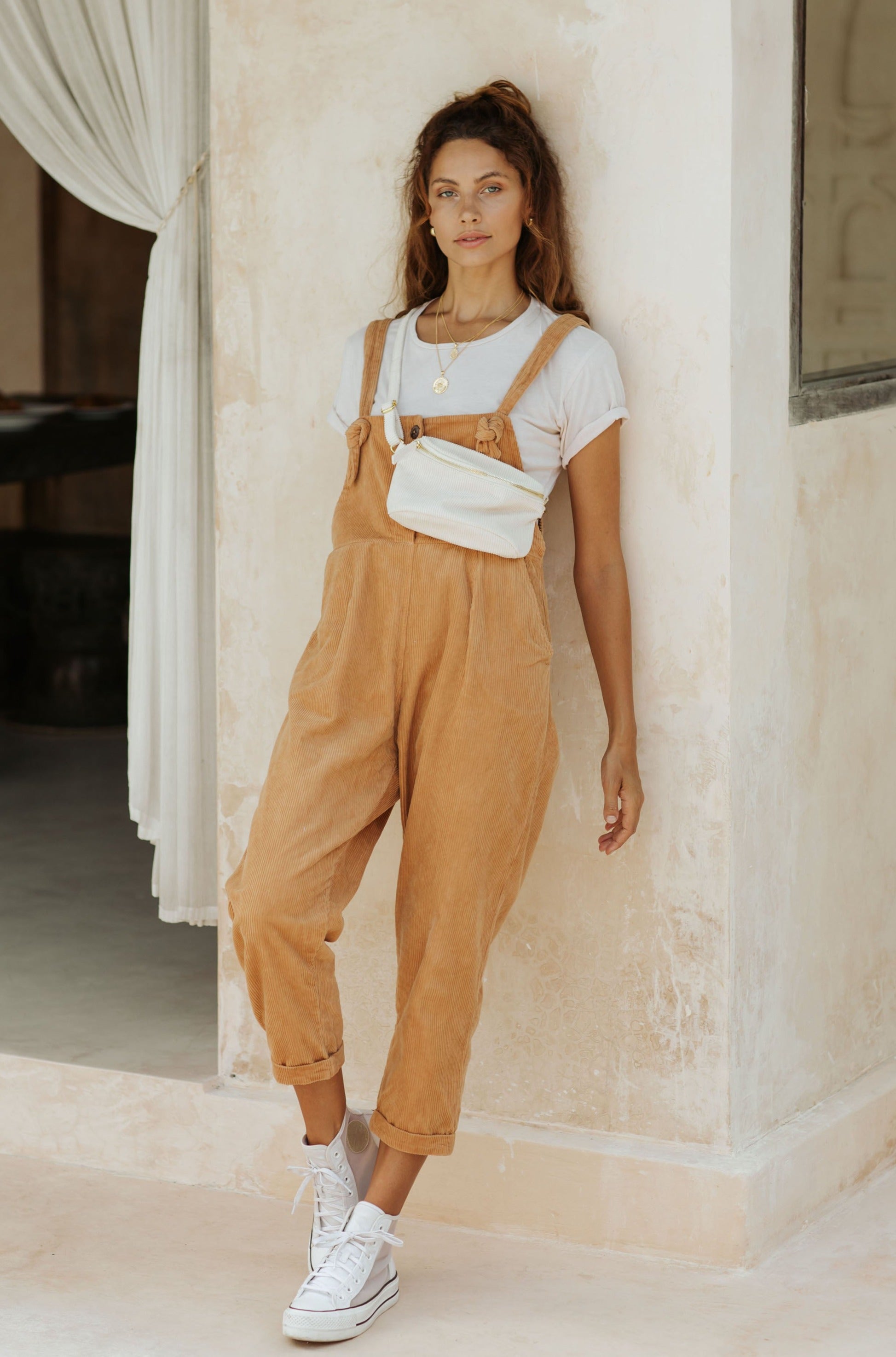 A woman in tan overalls and a white t-shirt, wearing a white fanny pack, standing against a wall with white curtains in the background.