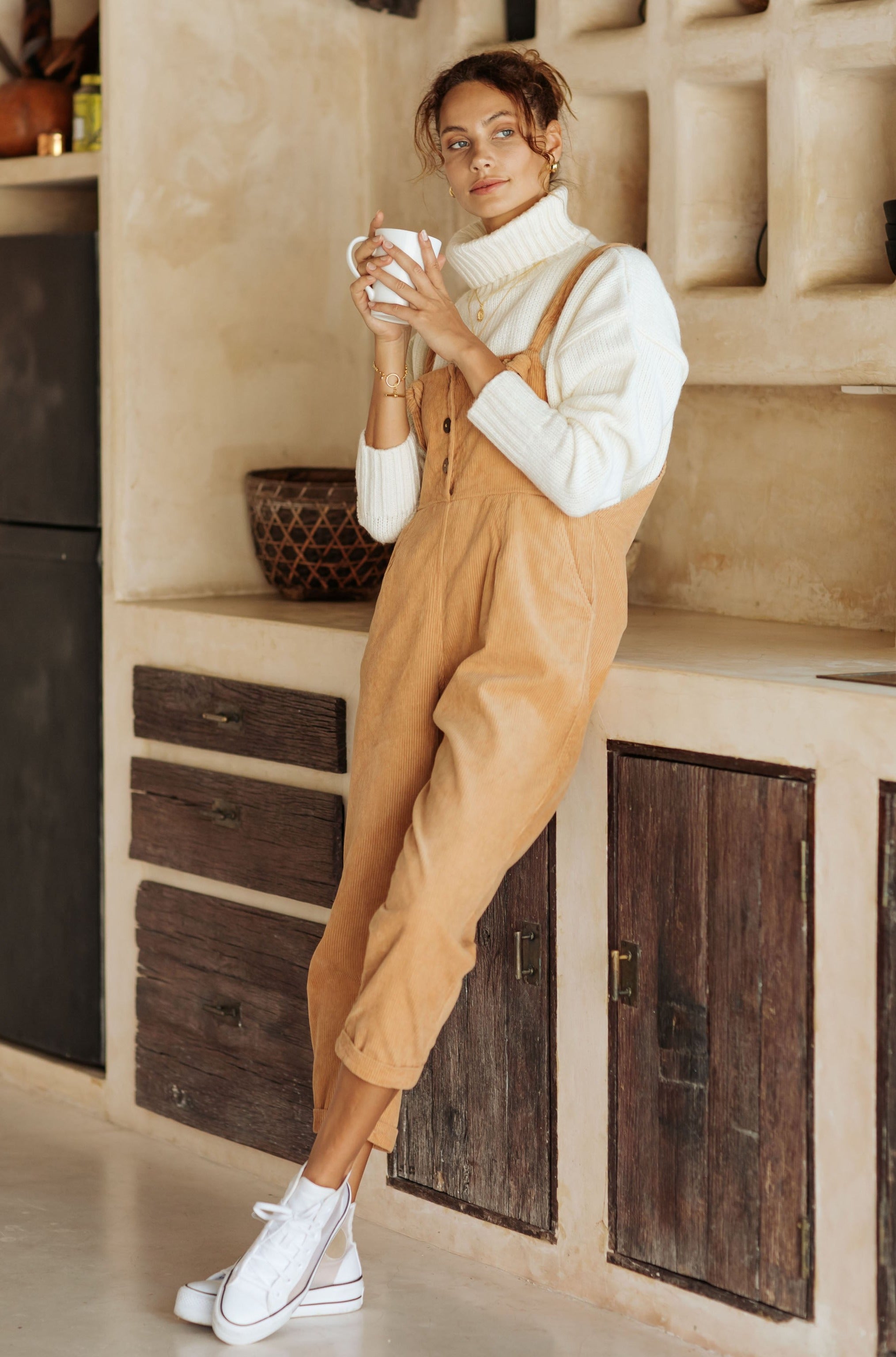 A woman in tan overalls and a white turtleneck, holding a mug while leaning against a kitchen counter.