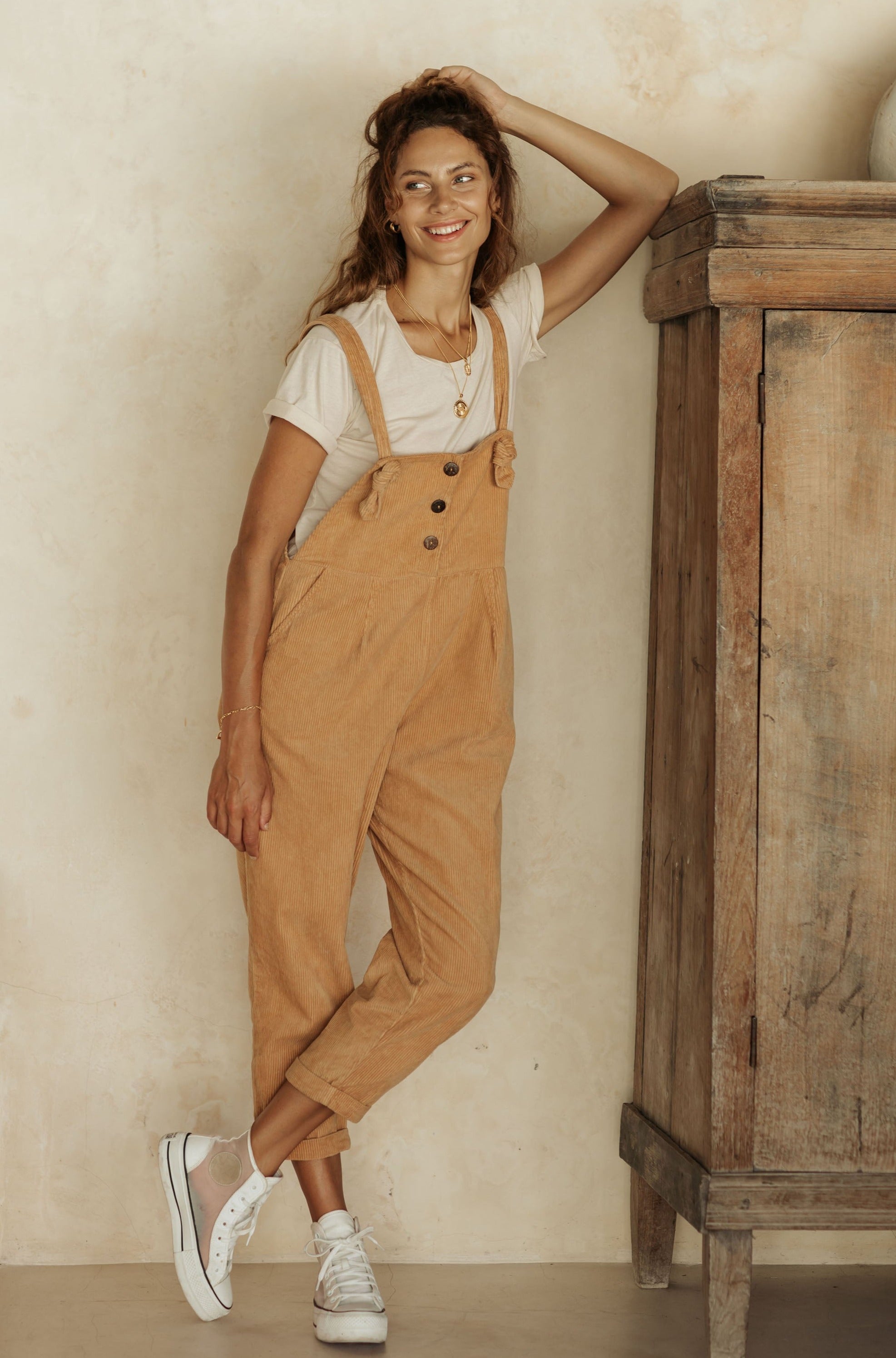 A woman wearing tan overalls and a white t-shirt, leaning against a wall with a wooden cabinet beside her.
