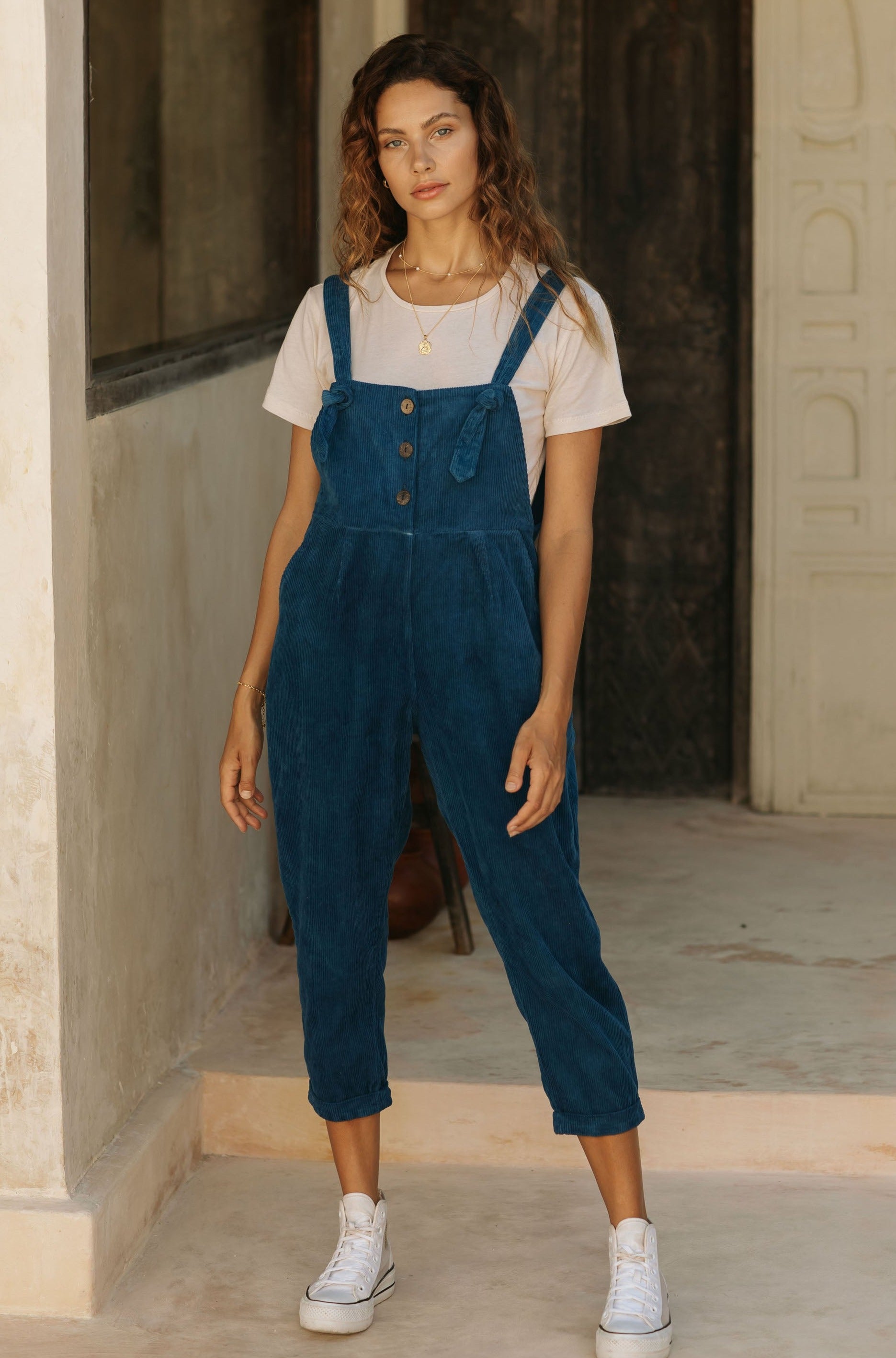 A woman in blue overalls and a white t-shirt, standing in a rustic setting.