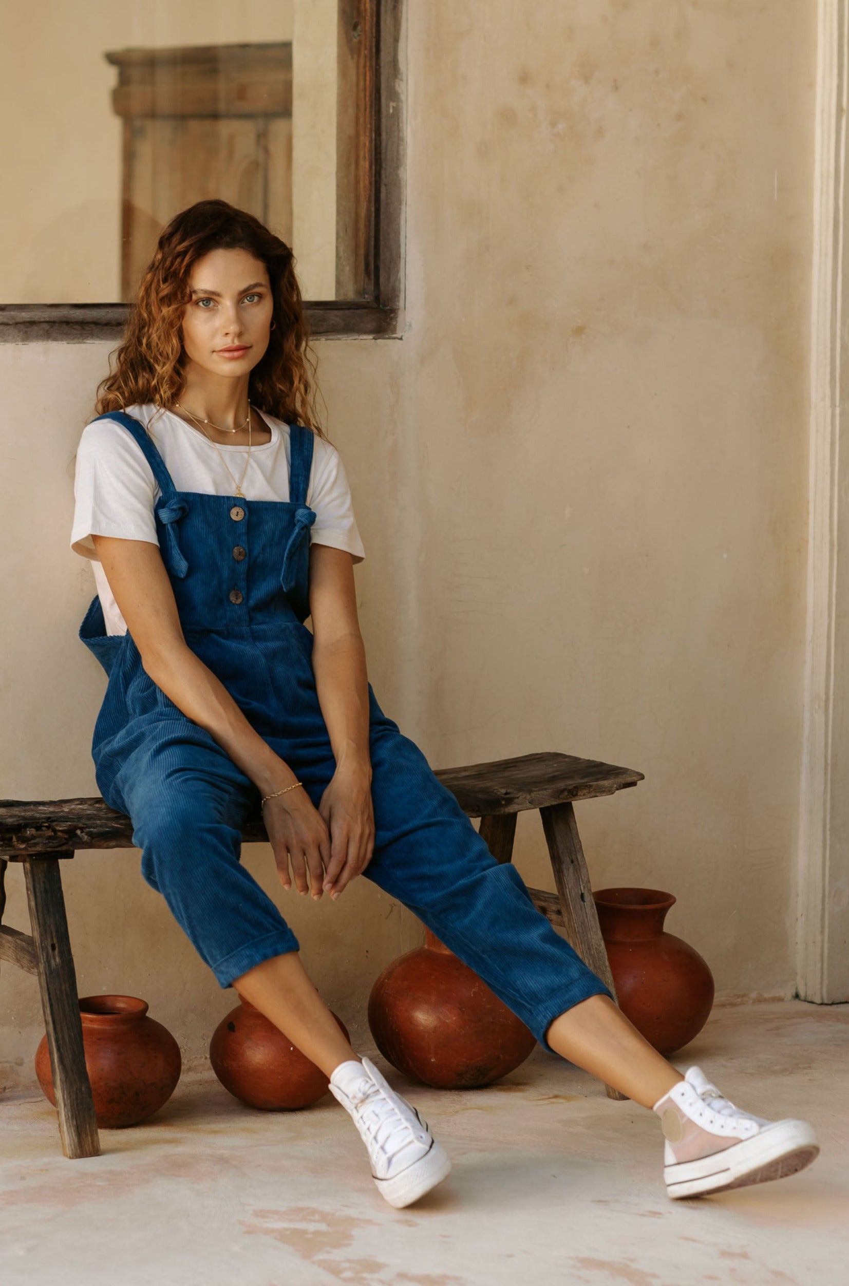A woman in blue overalls and a white t-shirt, sitting on a wooden bench with clay pots underneath.