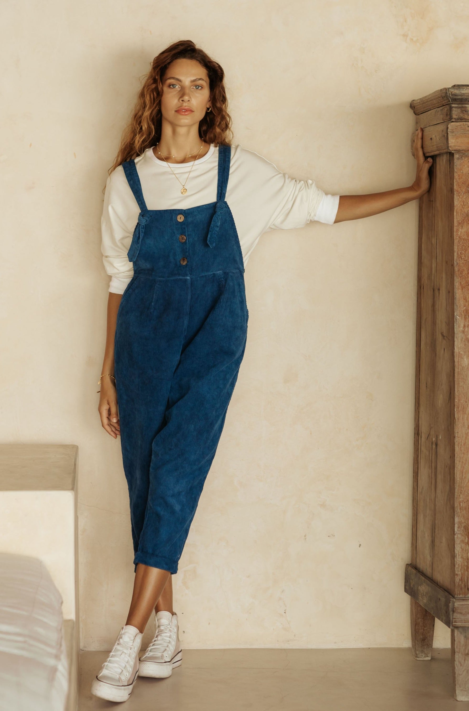 A woman in blue overalls and a white long-sleeve top, leaning against a wooden cabinet