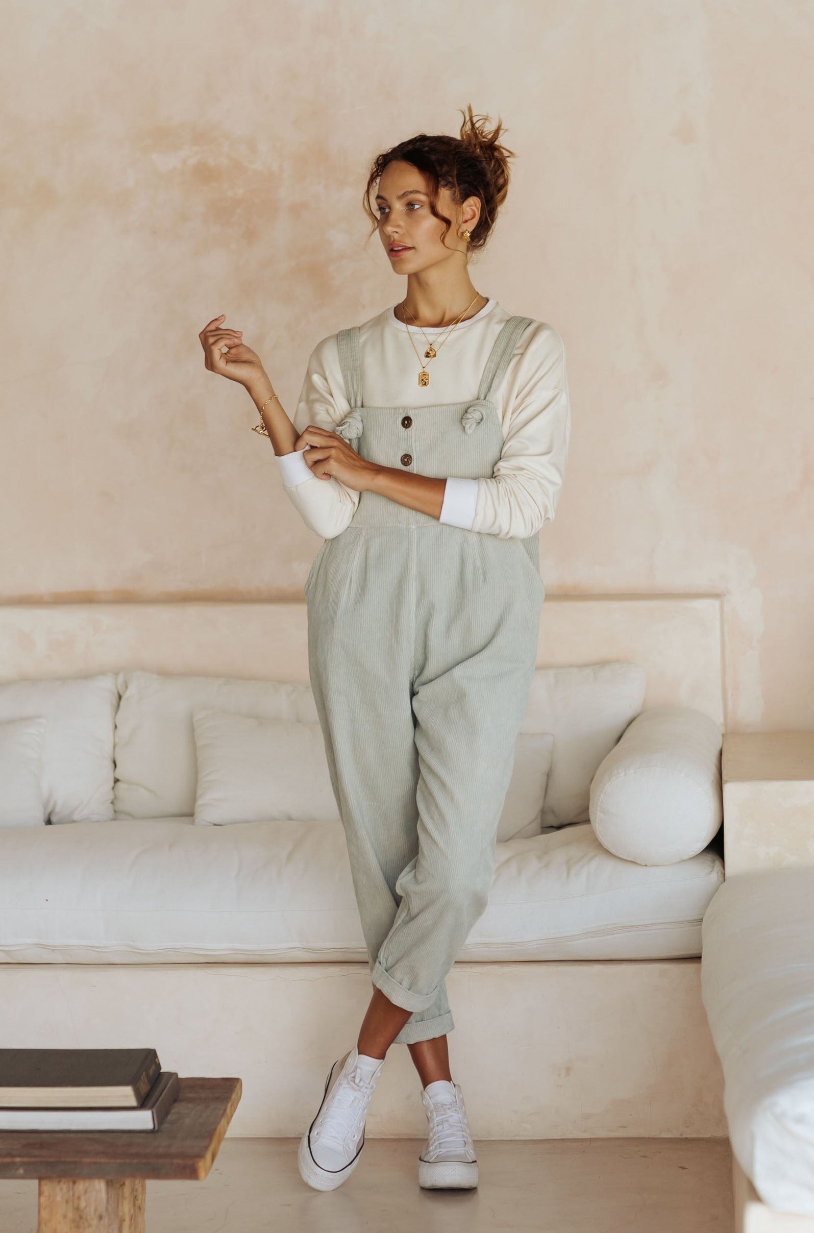 A woman in light green overalls and a white long-sleeve top, standing in a room with a white couch and neutral decor.