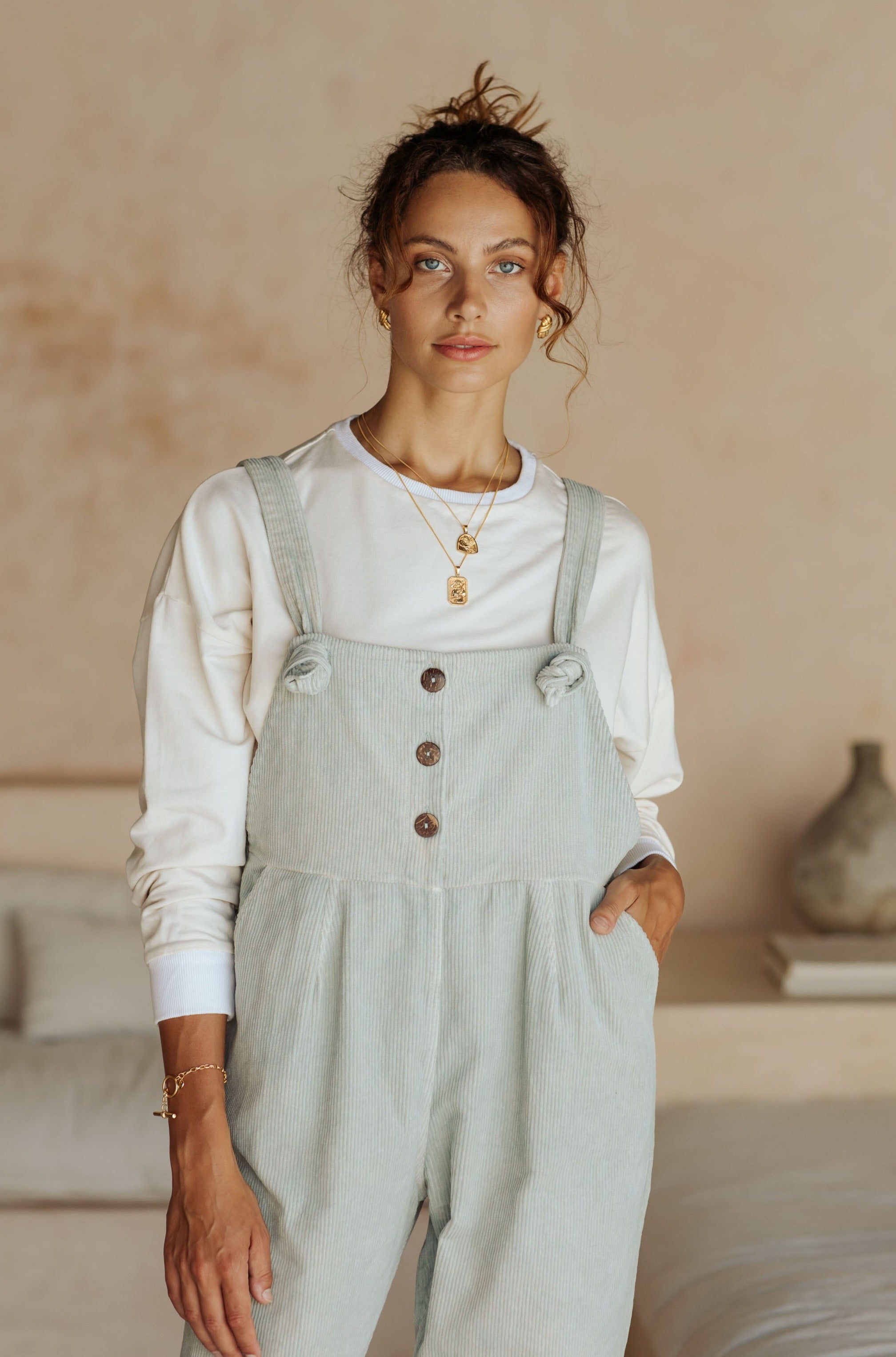 A woman in light green overalls and a white long-sleeve top, standing in a room with neutral decor and looking at the camera.