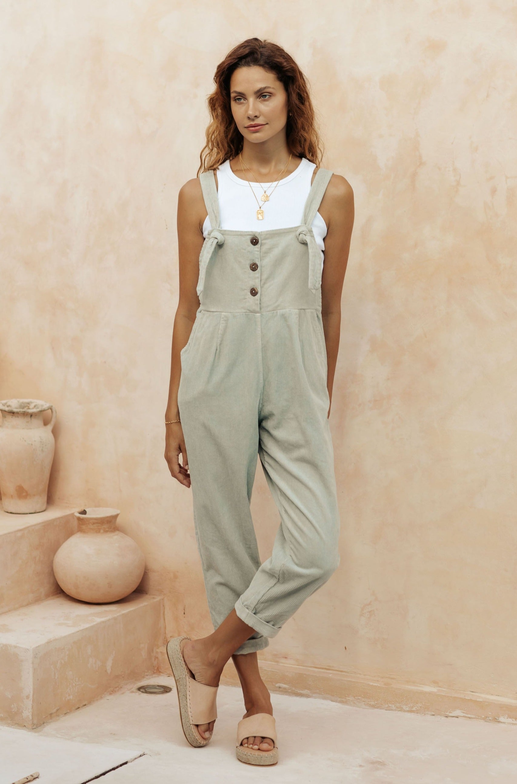A woman in light green overalls and a white tank top, standing in front of a neutral-toned wall with clay pots in the background.