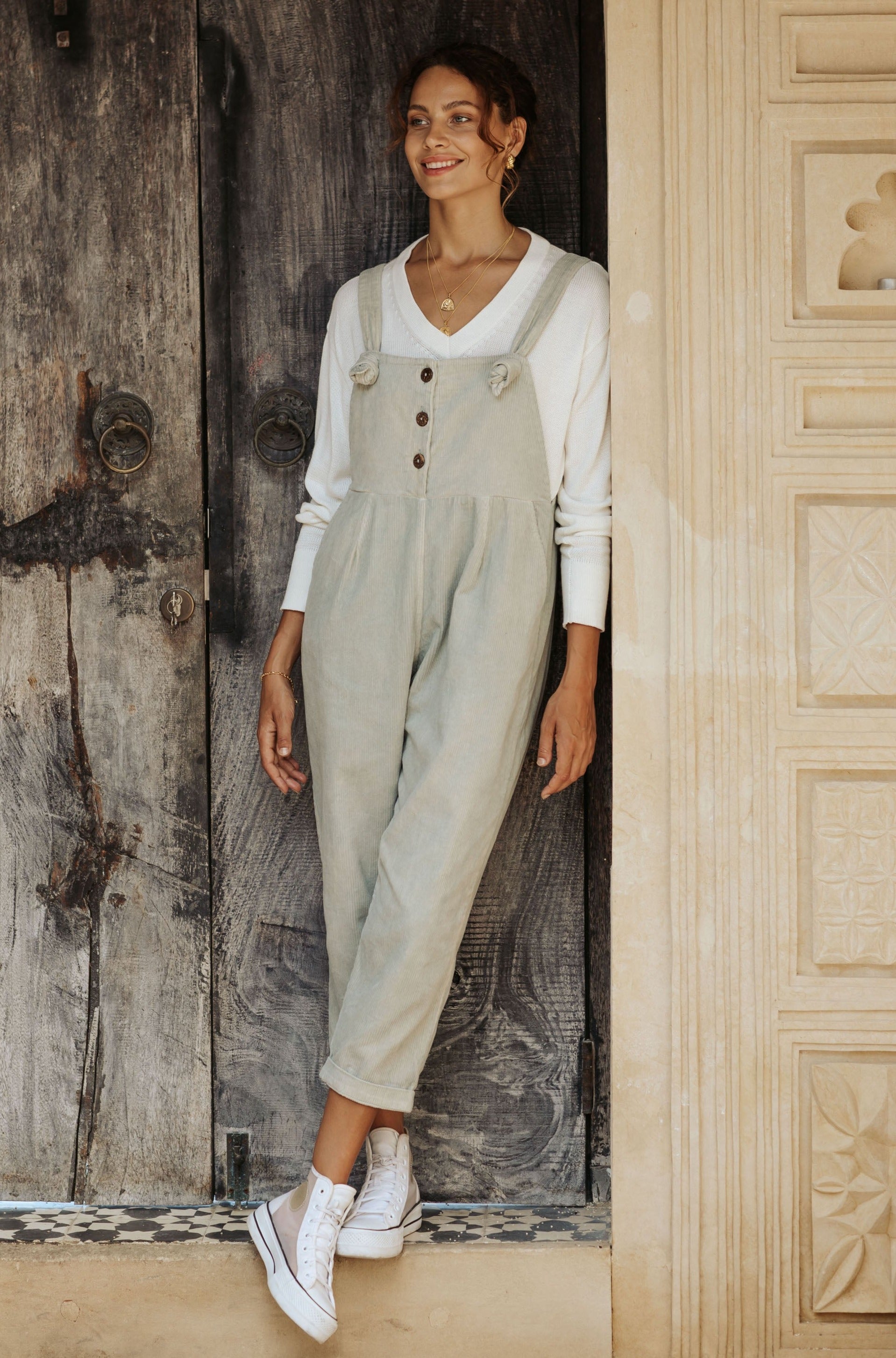 A woman in sage overalls and a white tank top, wearing white sneakers leans against a decorative wall.