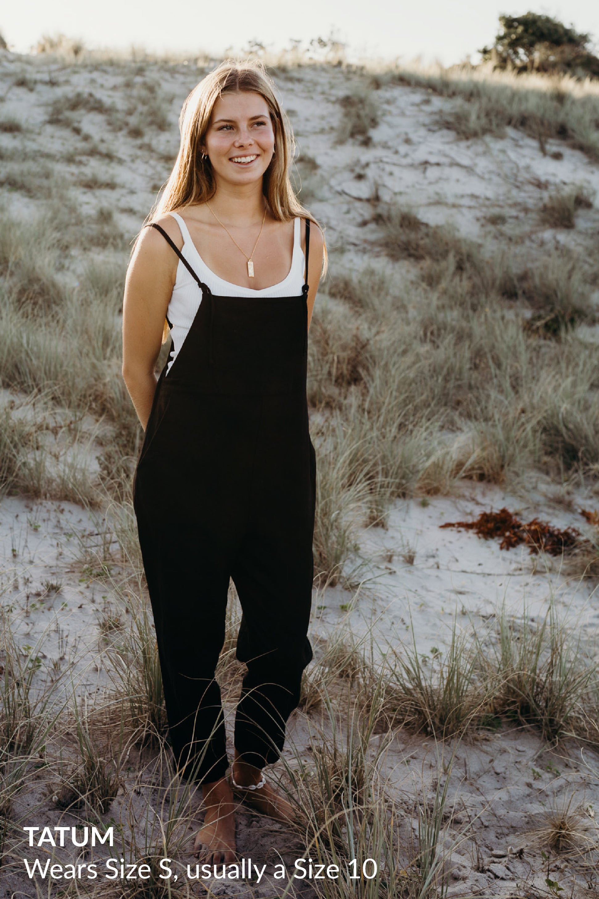 A woman smiling and standing on grassy dunes wearing a white tank top under black overalls and gold jewelry.