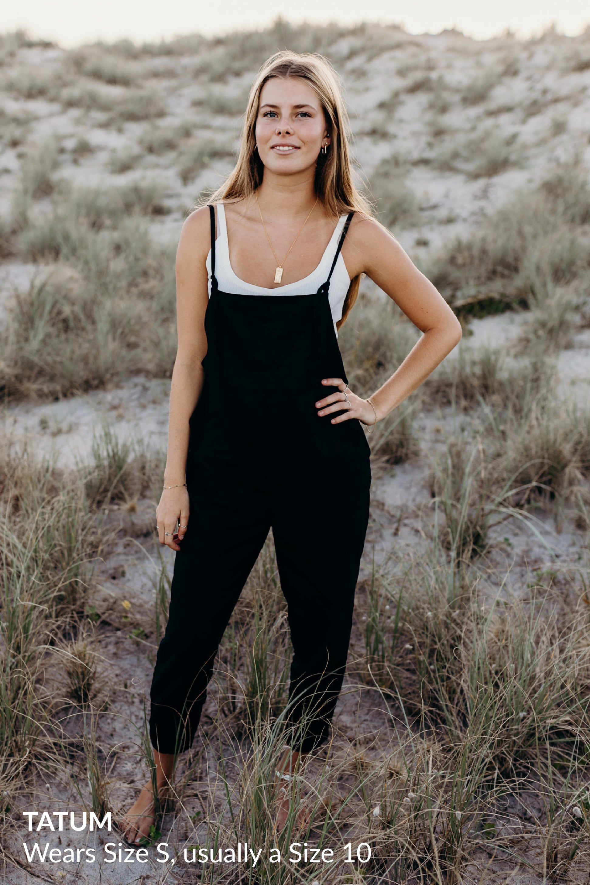 A woman standing on grassy dunes wearing a white tank top under black overalls and gold jewelry.