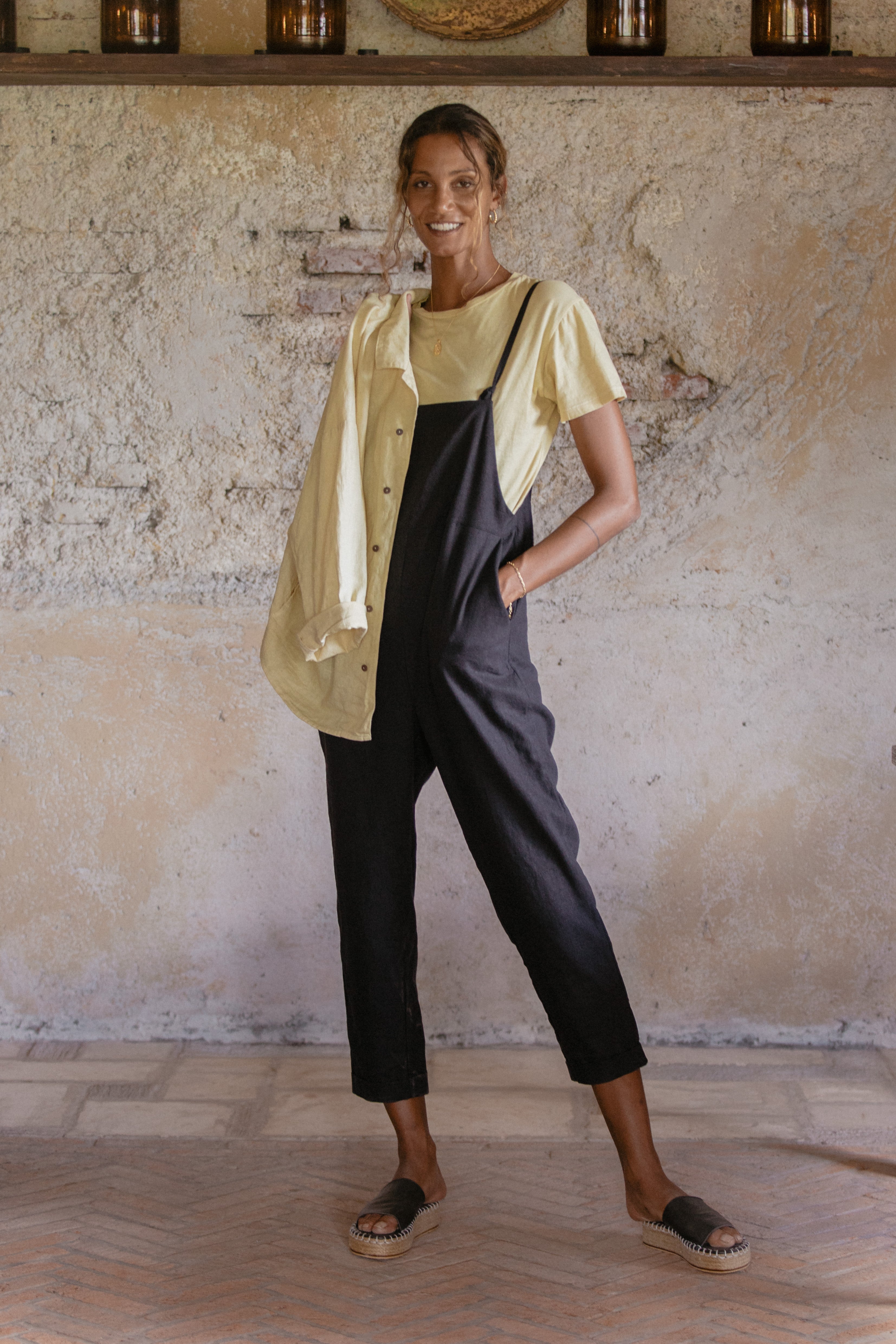 A woman standing indoors, smiling and wearing a yellow t-shirt under black overalls with black espadrille sandals.