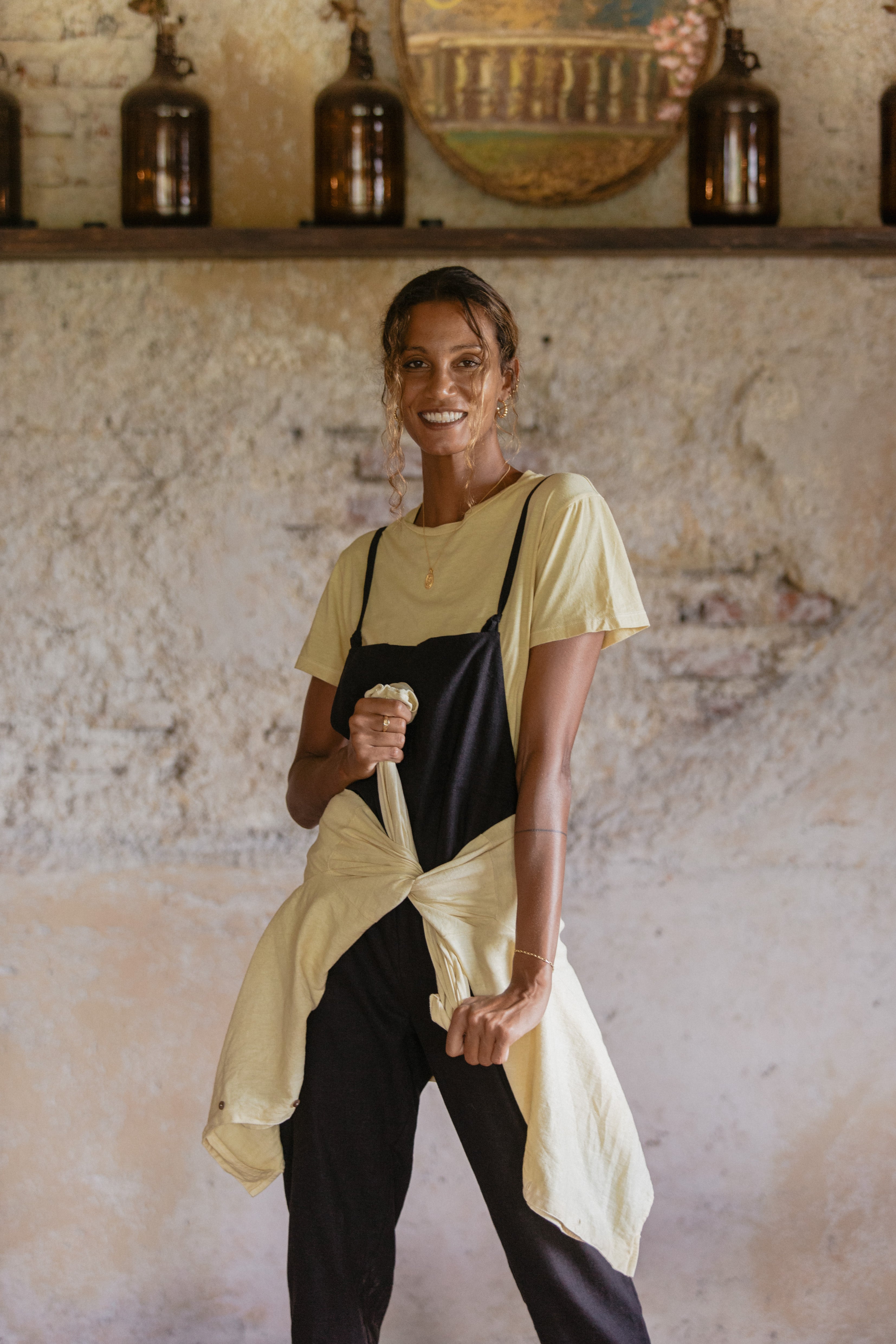 A woman smiling indoors, wearing a yellow t-shirt under black overalls with a tied yellow shirt around her waist.