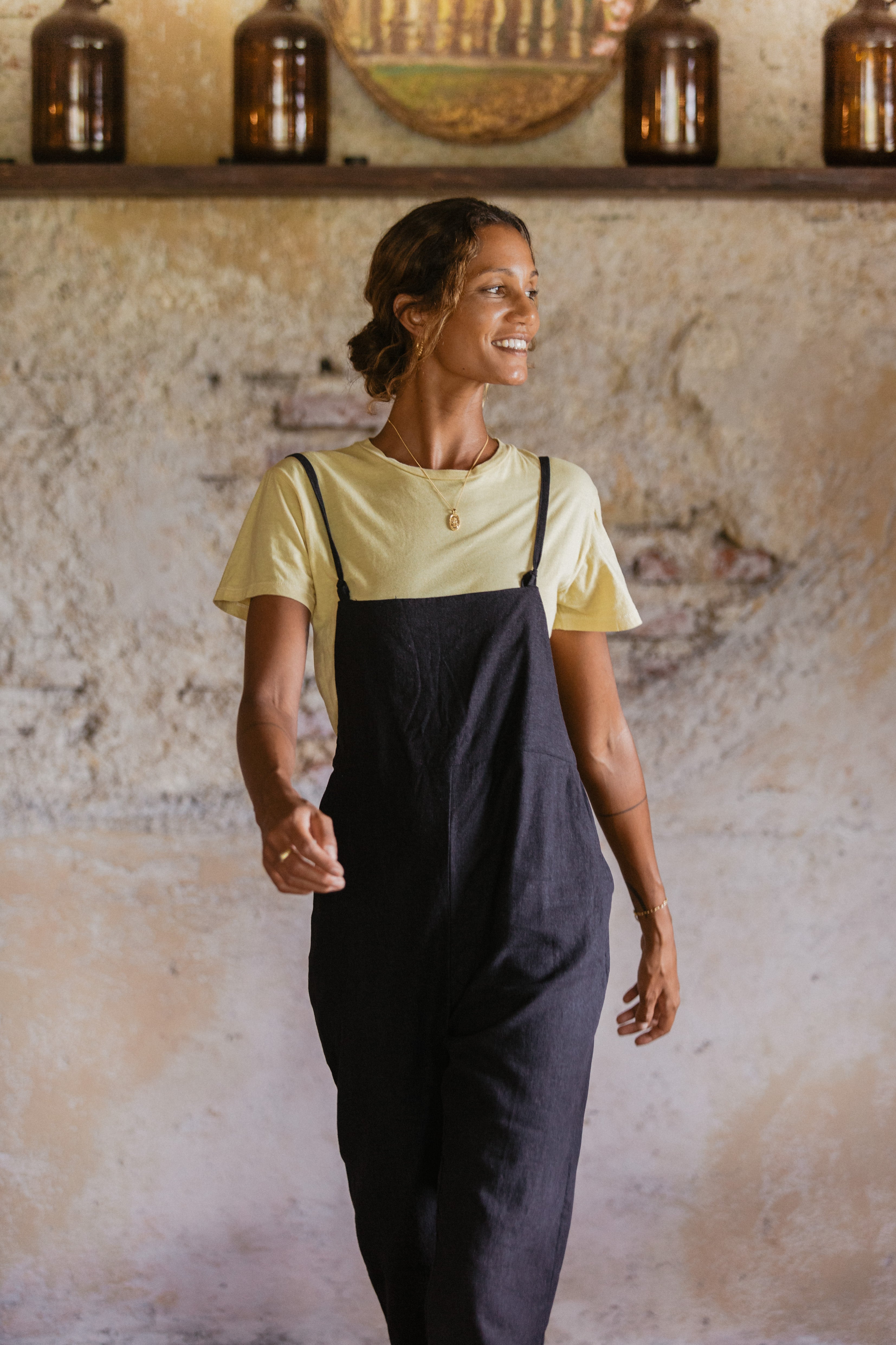 A woman smiling indoors and looking to the side, wearing a yellow t-shirt under black overalls.