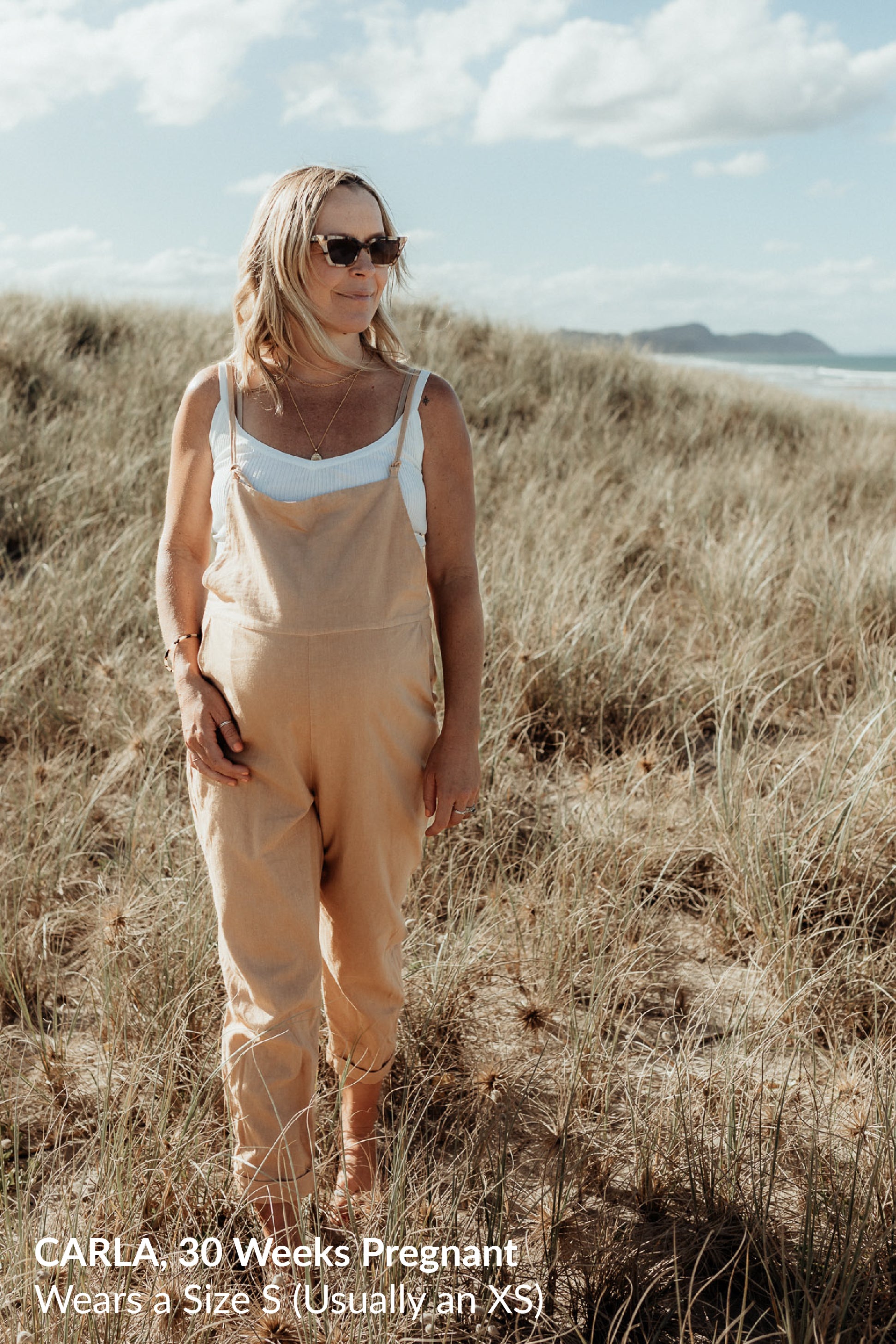 A pregnant woman wearing sunglasses, beige overalls, and a white top, standing on grassy dunes