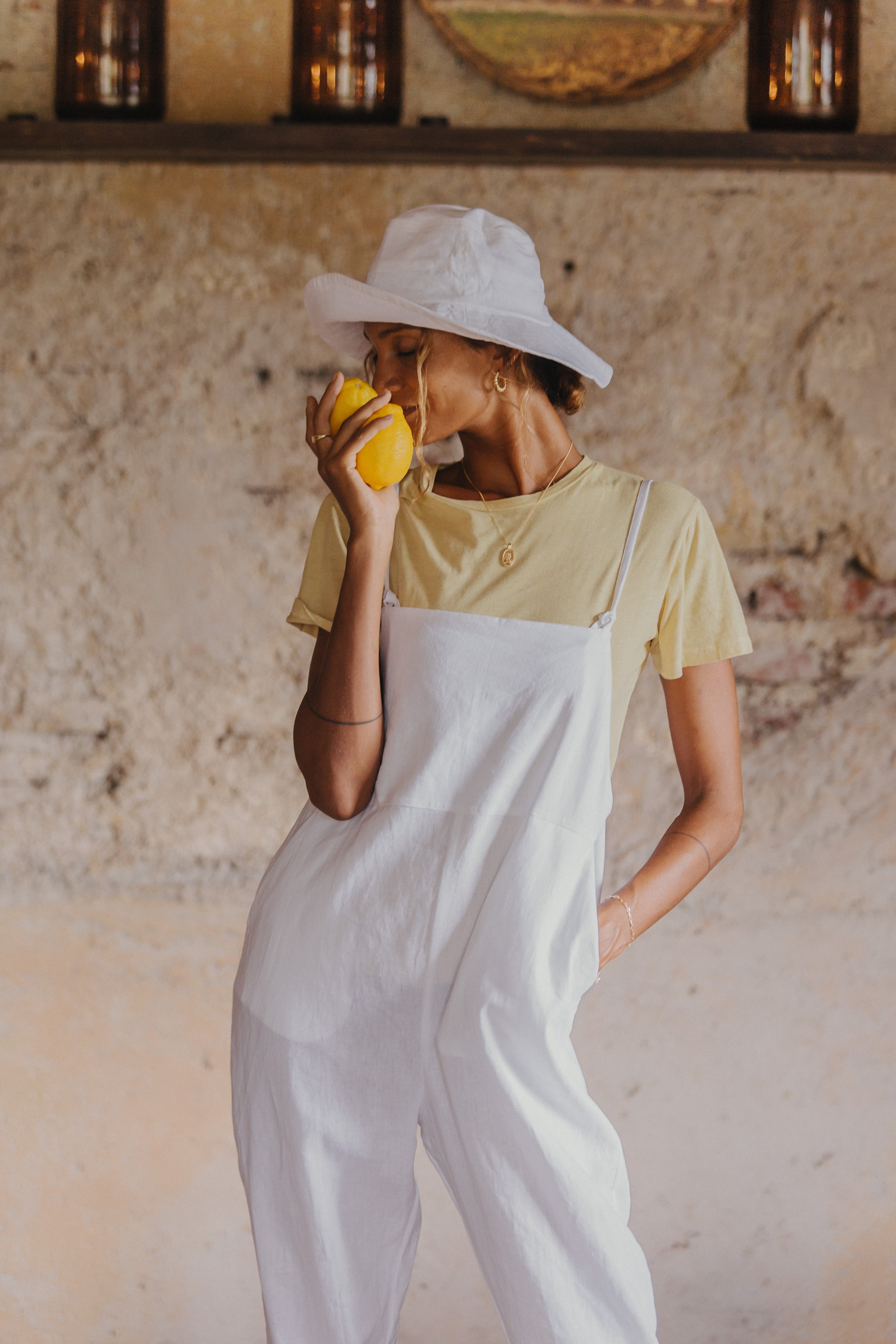 A woman wearing a white hat, yellow t-shirt, and white overalls, holds a lemon close to her face.