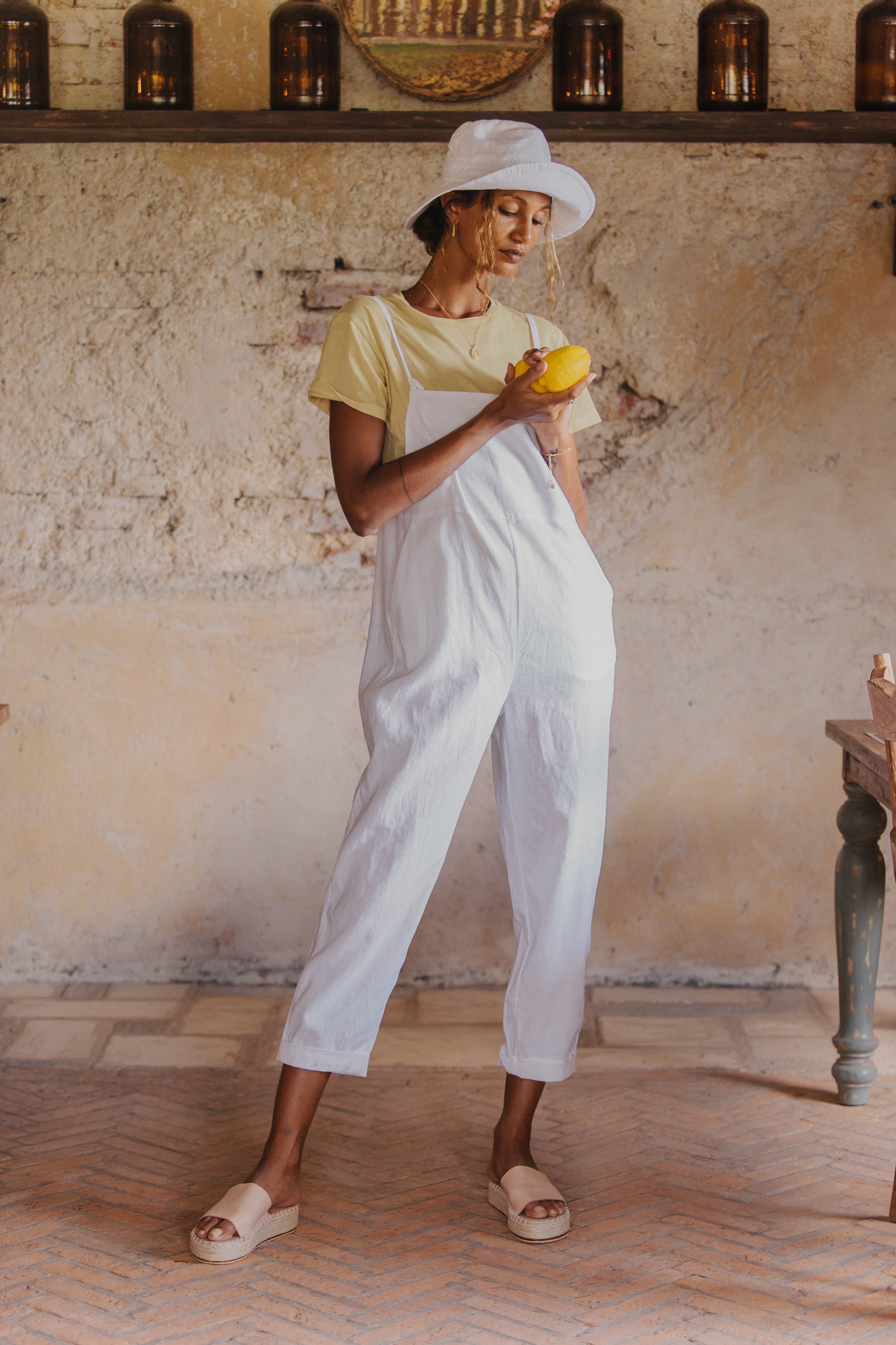 A woman wearing a white hat, yellow t-shirt, and white overalls, stands indoors holding a lemon with one hand in her pocket.