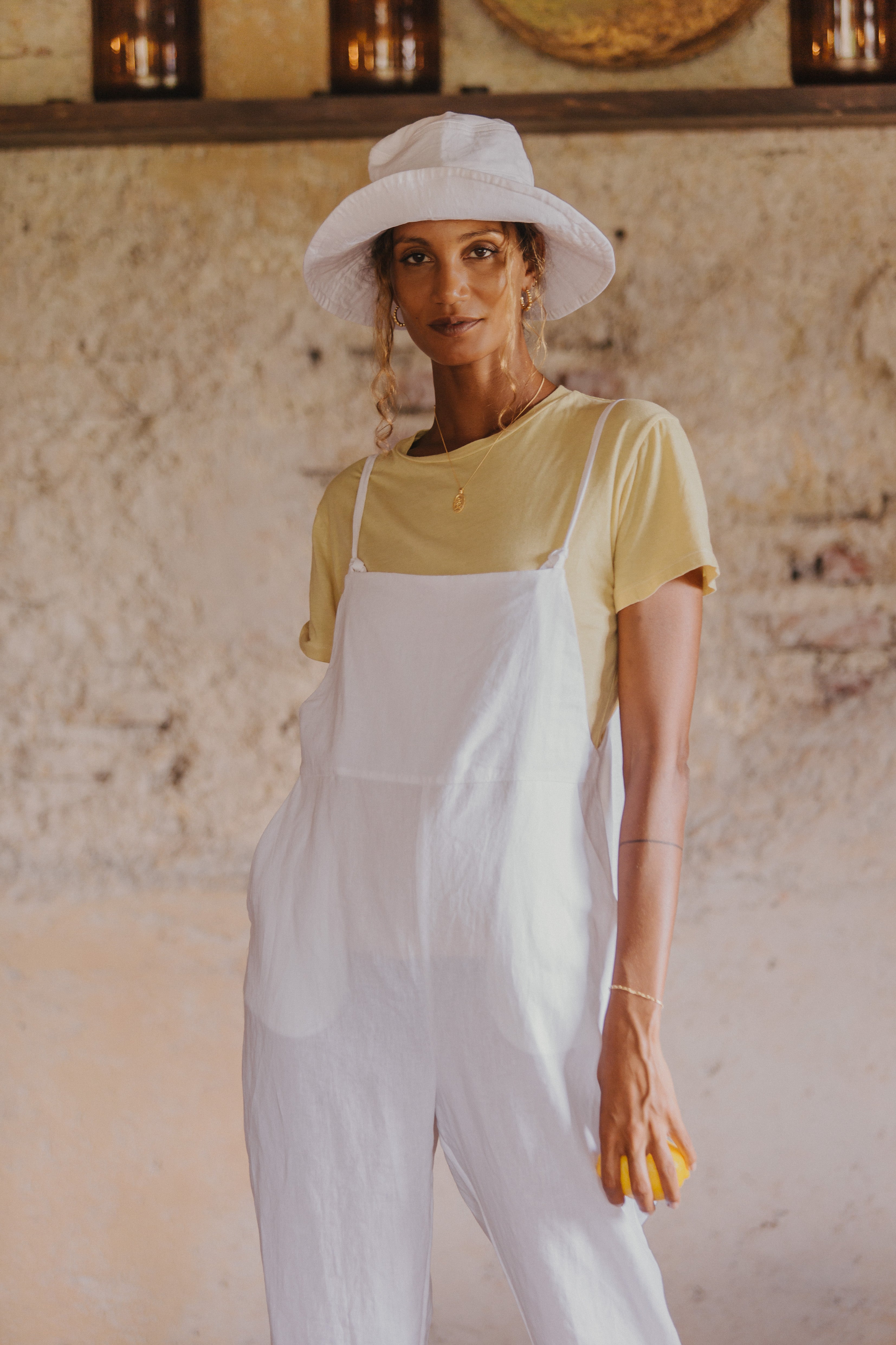 A woman wearing a white hat, yellow t-shirt, and white overalls, stands indoors holding a lemon.