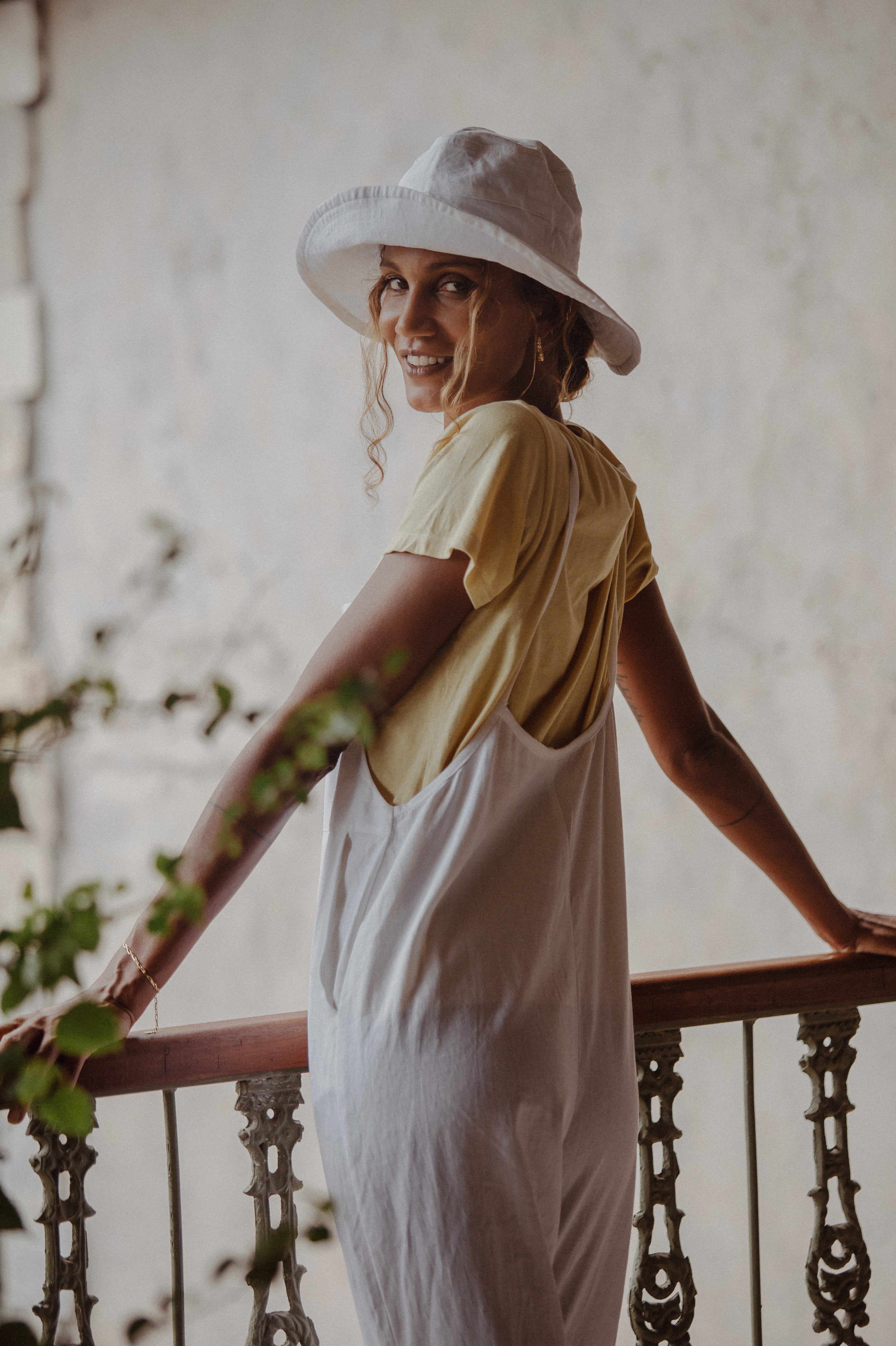 A woman standing on a balcony, wearing a white hat, yellow t-shirt, and white overalls, smiling over her shoulder.