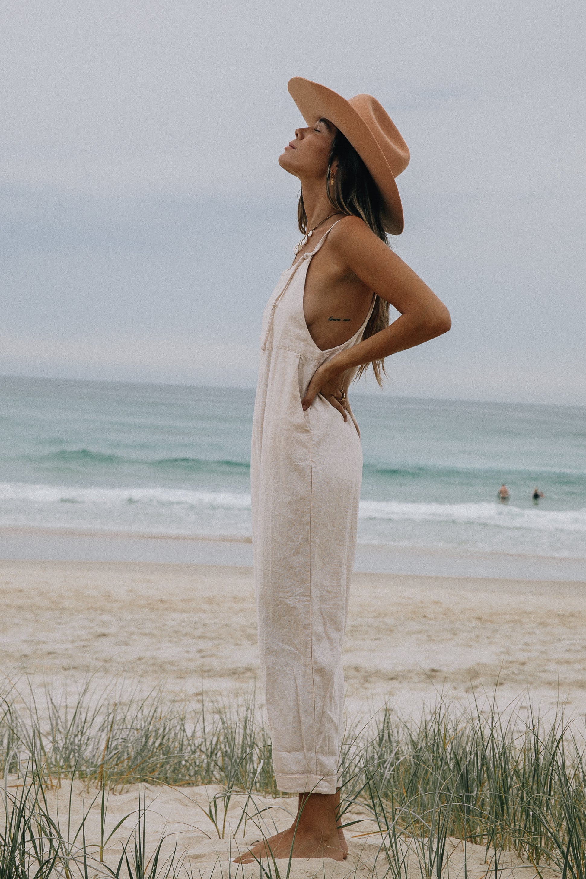 A side view of a woman standing on a beach in front of the ocean, wearing a wide-brimmed hat and light pink overalls over a tank top.