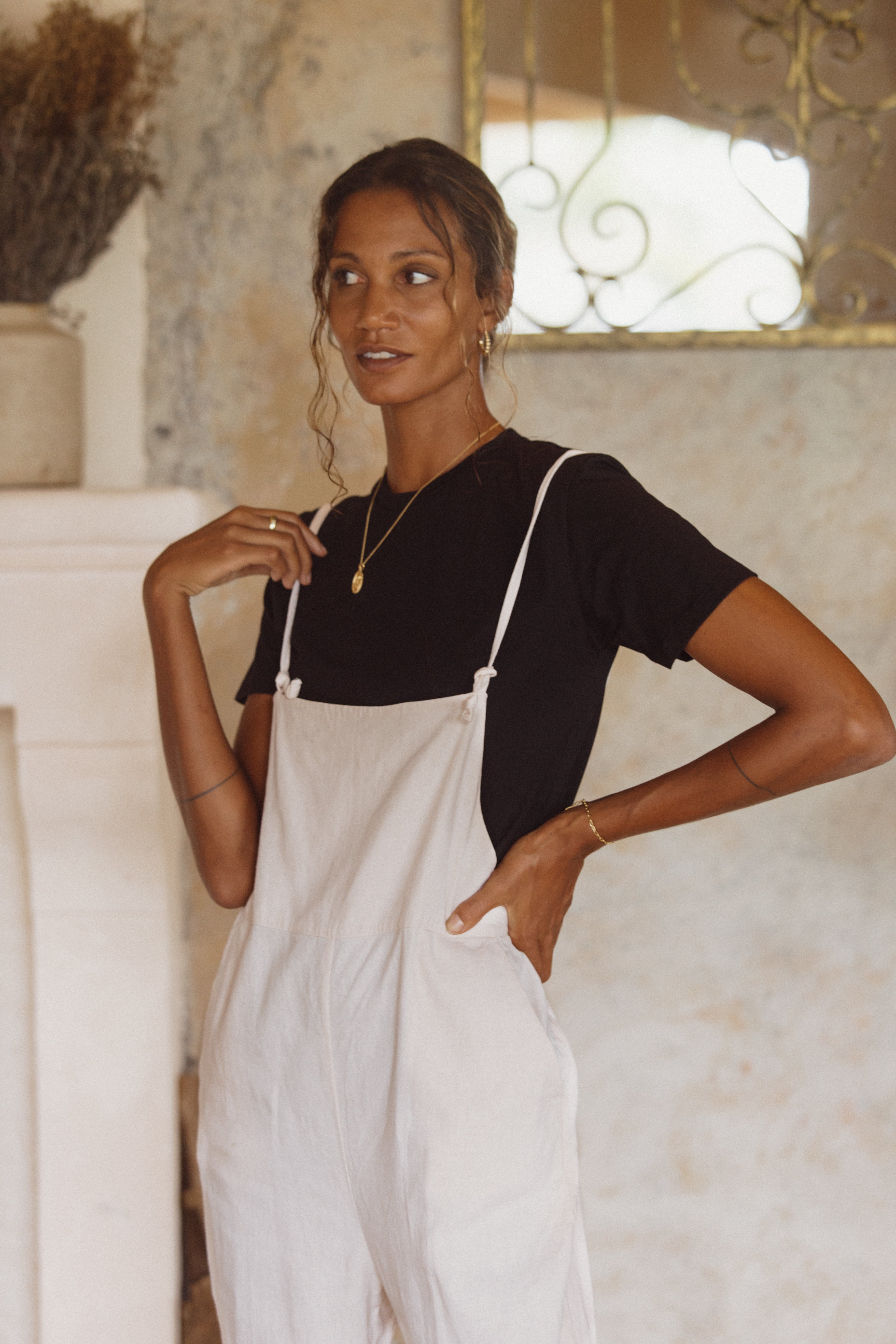 A woman wearing a black t-shirt and light pink overalls, standing indoors with one hand in her pocket.
