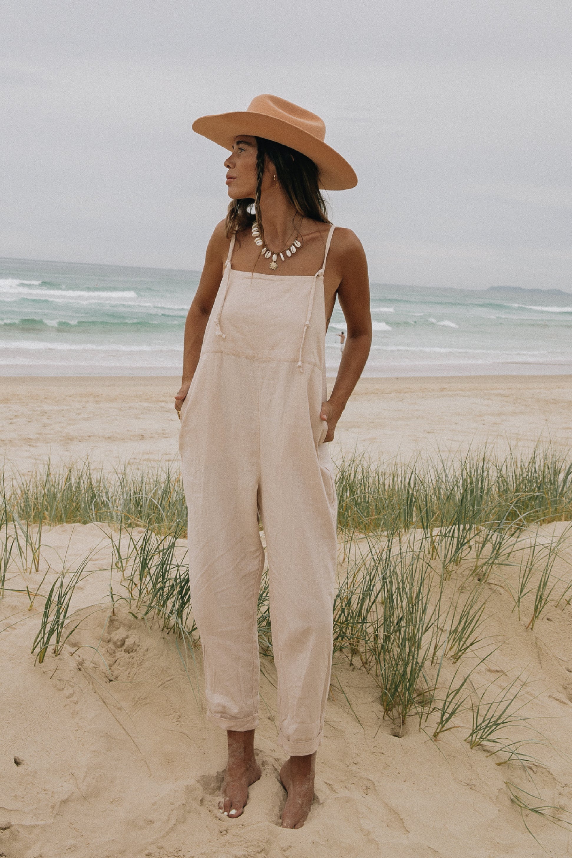A woman standing on a beach in front of the ocean, wearing a wide-brimmed hat, necklace, and light pink overalls over a tank top.