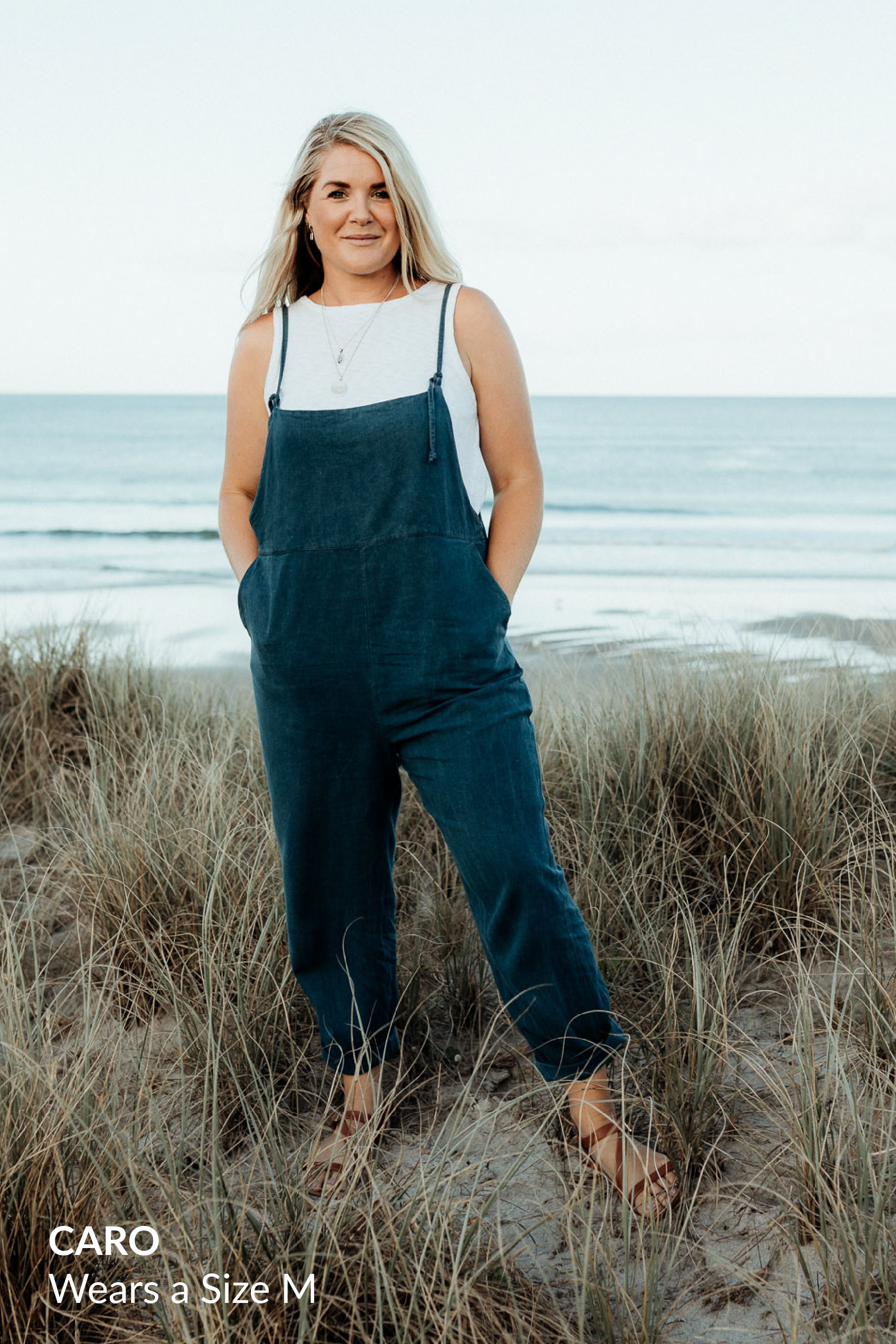 A blonde woman wearing a white tank top and dark blue overalls, standing on the beach in front of the ocean.