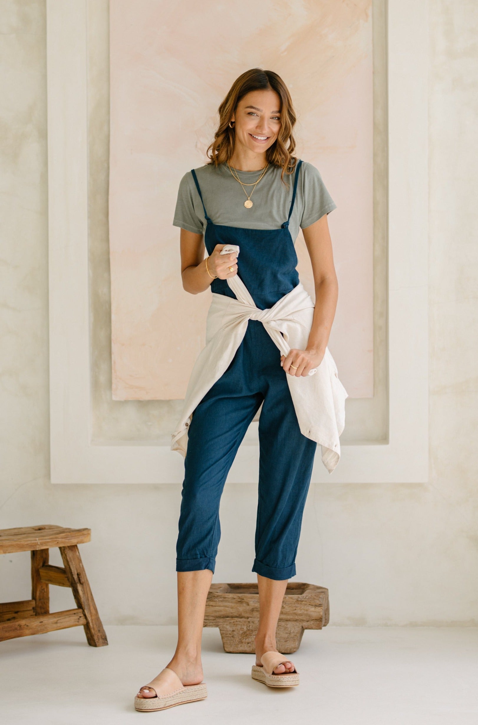 A woman wearing a gray t-shirt and dark blue overalls with a light jacket tied around her waist, standing indoors and smiling.