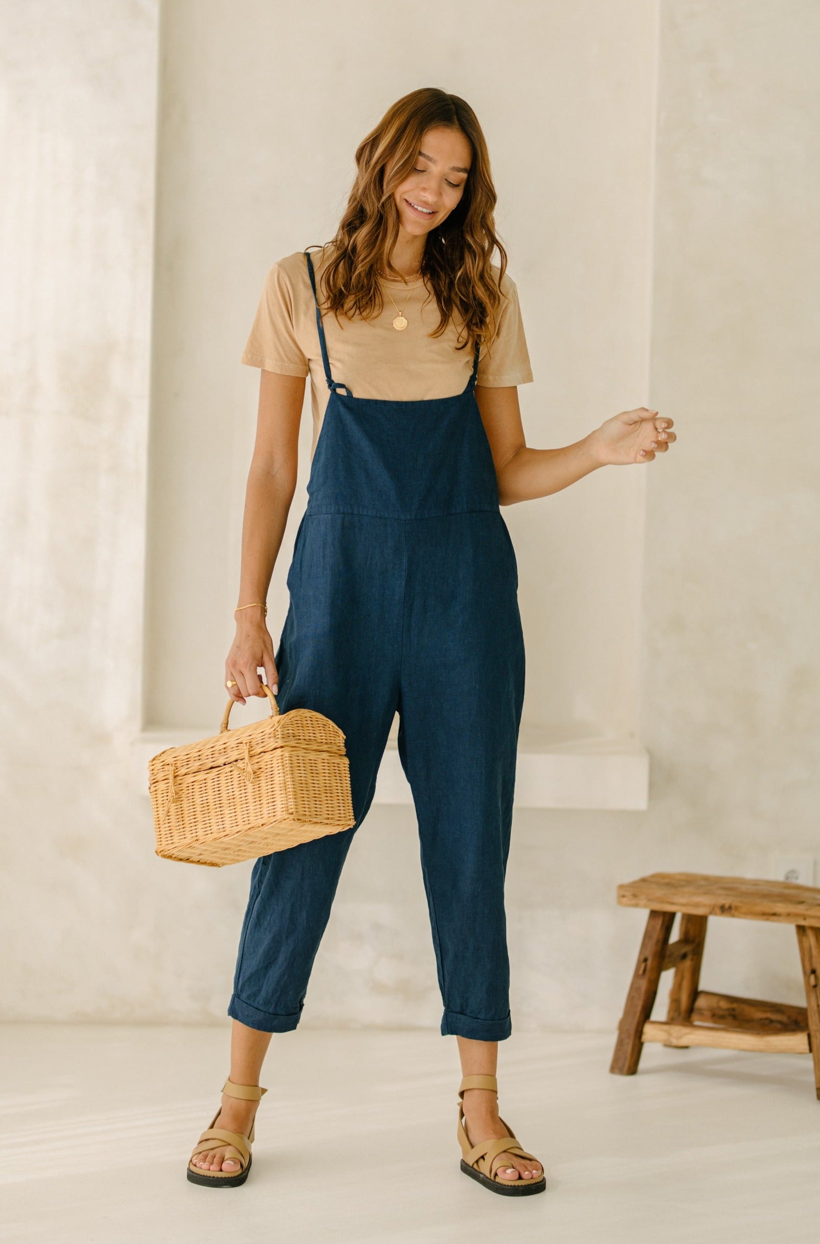 A woman wearing a beige t-shirt and dark blue overalls, holding a woven basket and looking down.