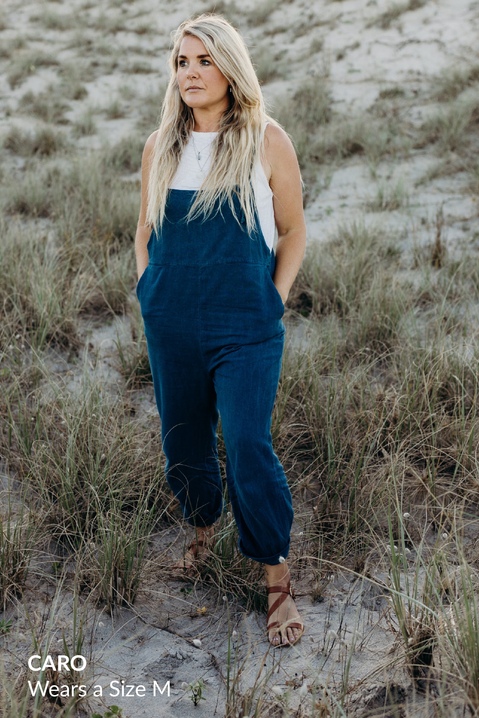 A blonde woman wearing a white tank top and dark blue overalls, standing on grassy dunes and looking to the side.