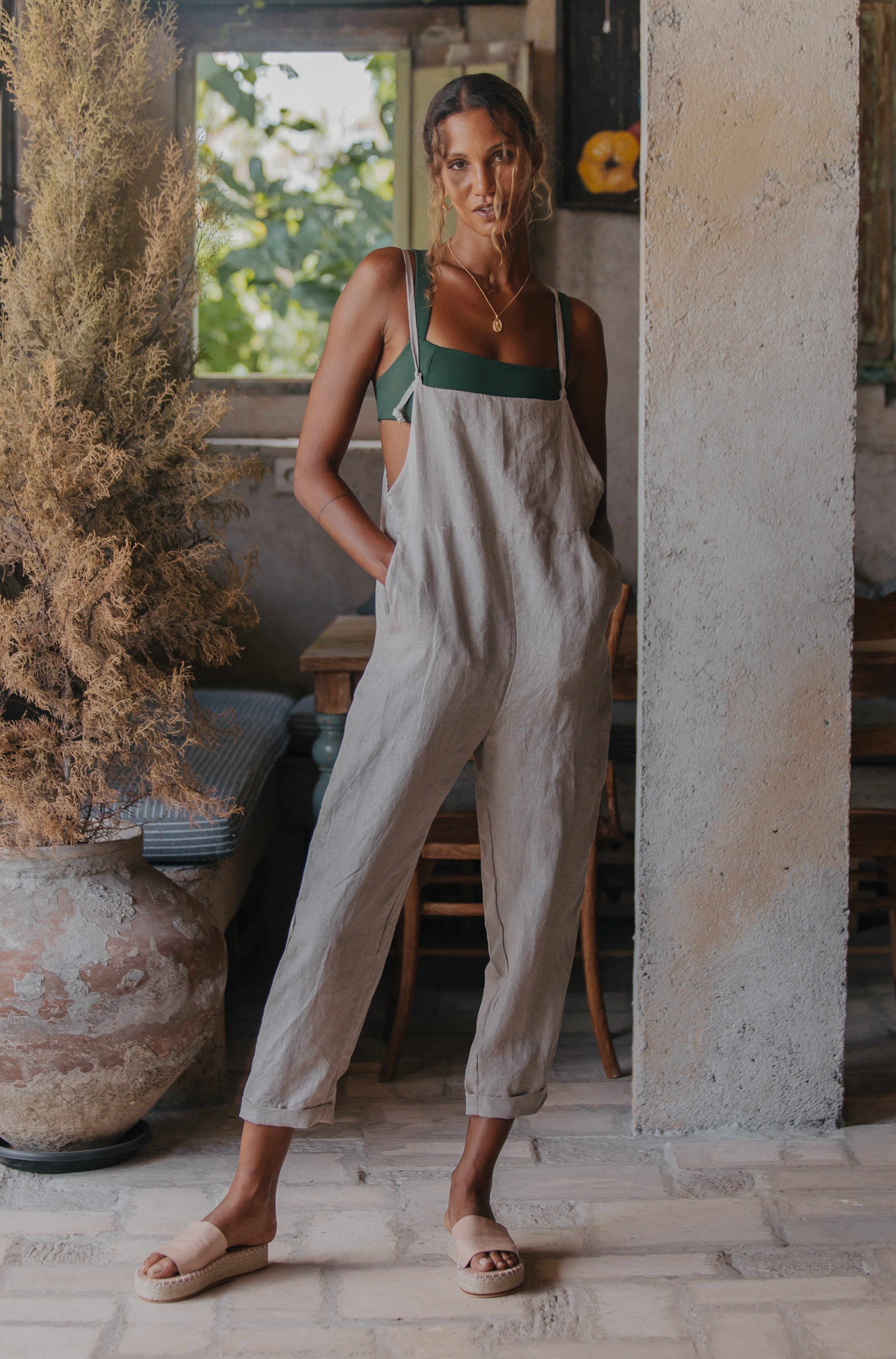 A woman in a slightly different pose but still wearing light beige overalls over a green top, standing indoors.