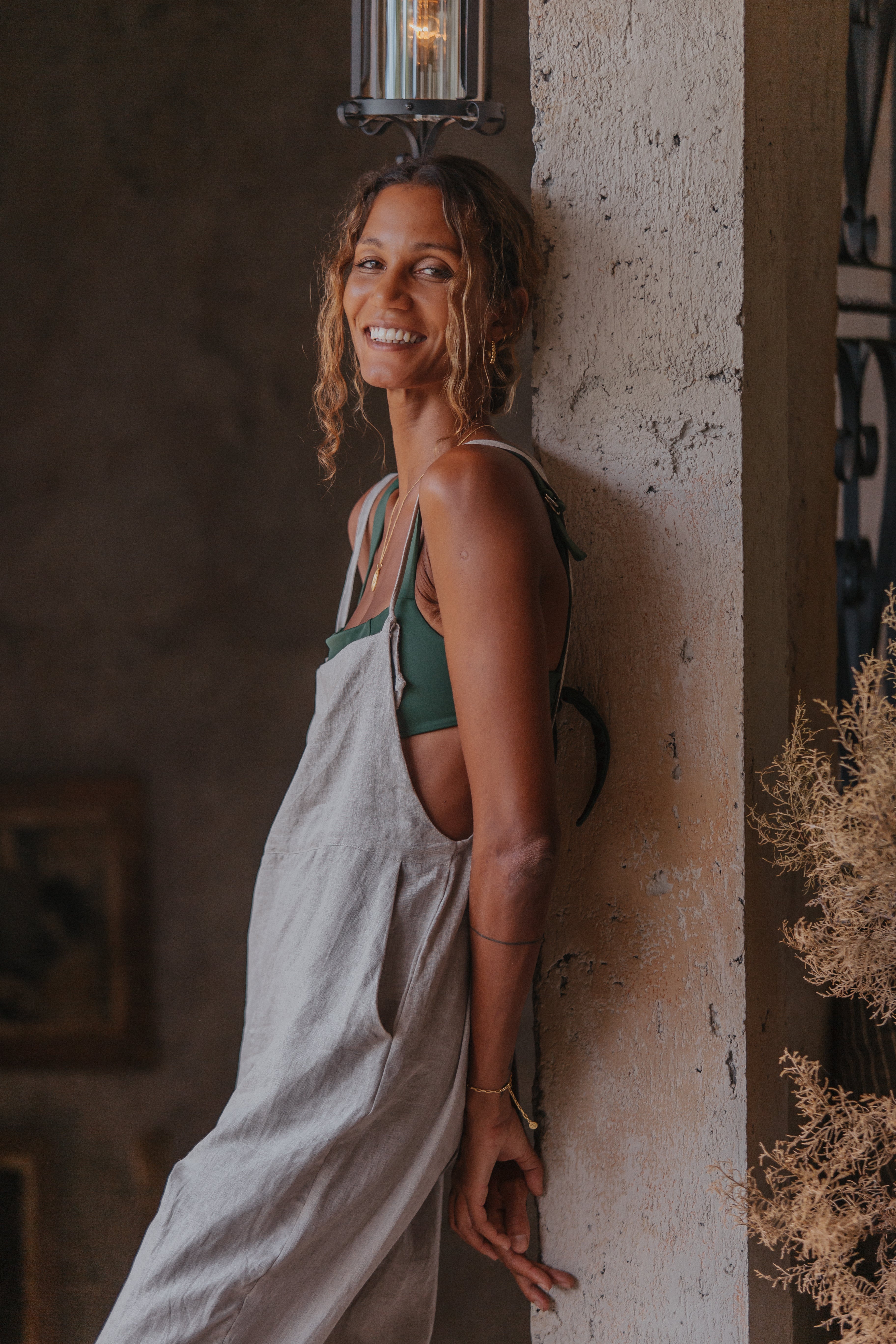 A woman wearing light beige overalls over a green top is smiling while leaning against a textured wall indoors, with a vintage-style lamp above her.