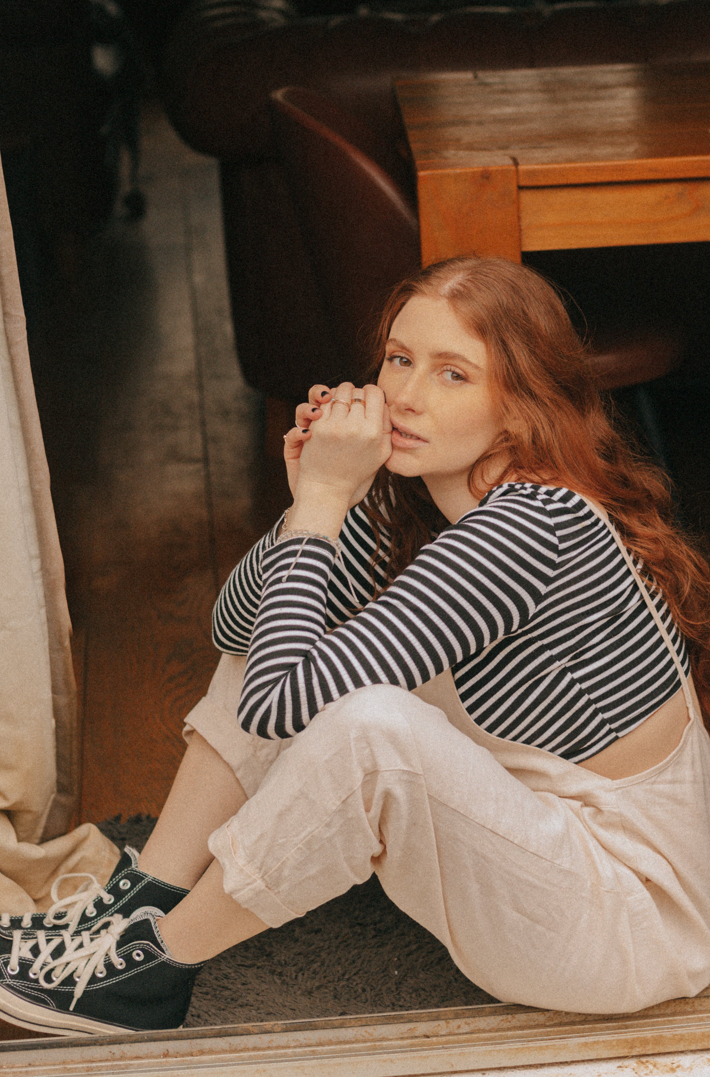 A woman with long red hair, sitting indoors near a door frame, wearing light beige overalls over a black and white striped long-sleeve top and black Converse sneakers.