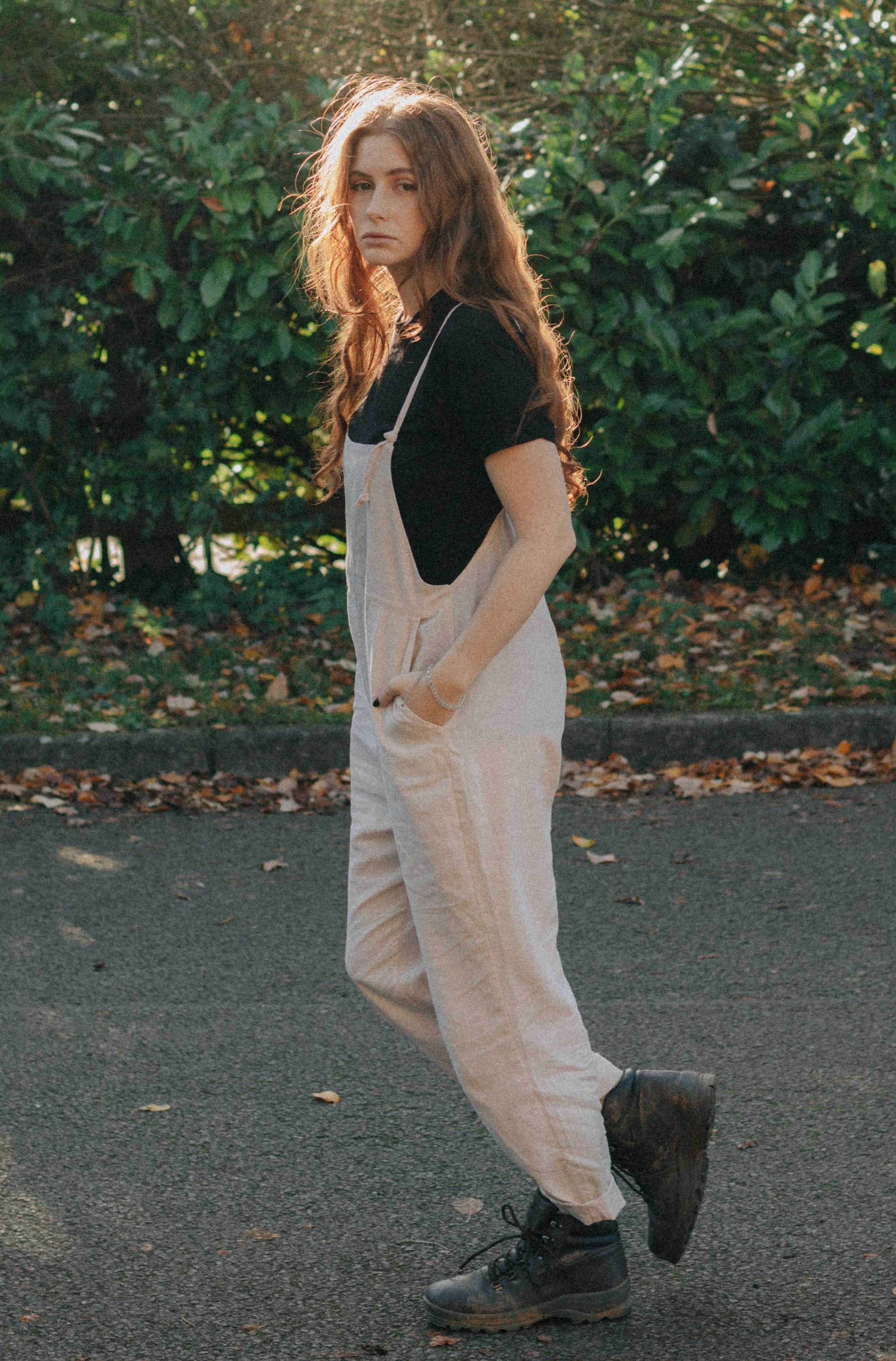 A woman with long, wavy red hair wearing light beige overalls and a black t-shirt, standing outdoors on a road with green bushes in the background.