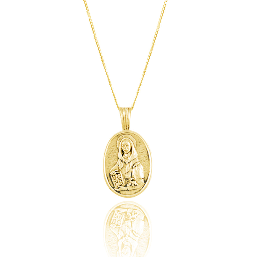 A gold oval medallion necklace featuring an engraved religious figure displayed on a reflective surface.