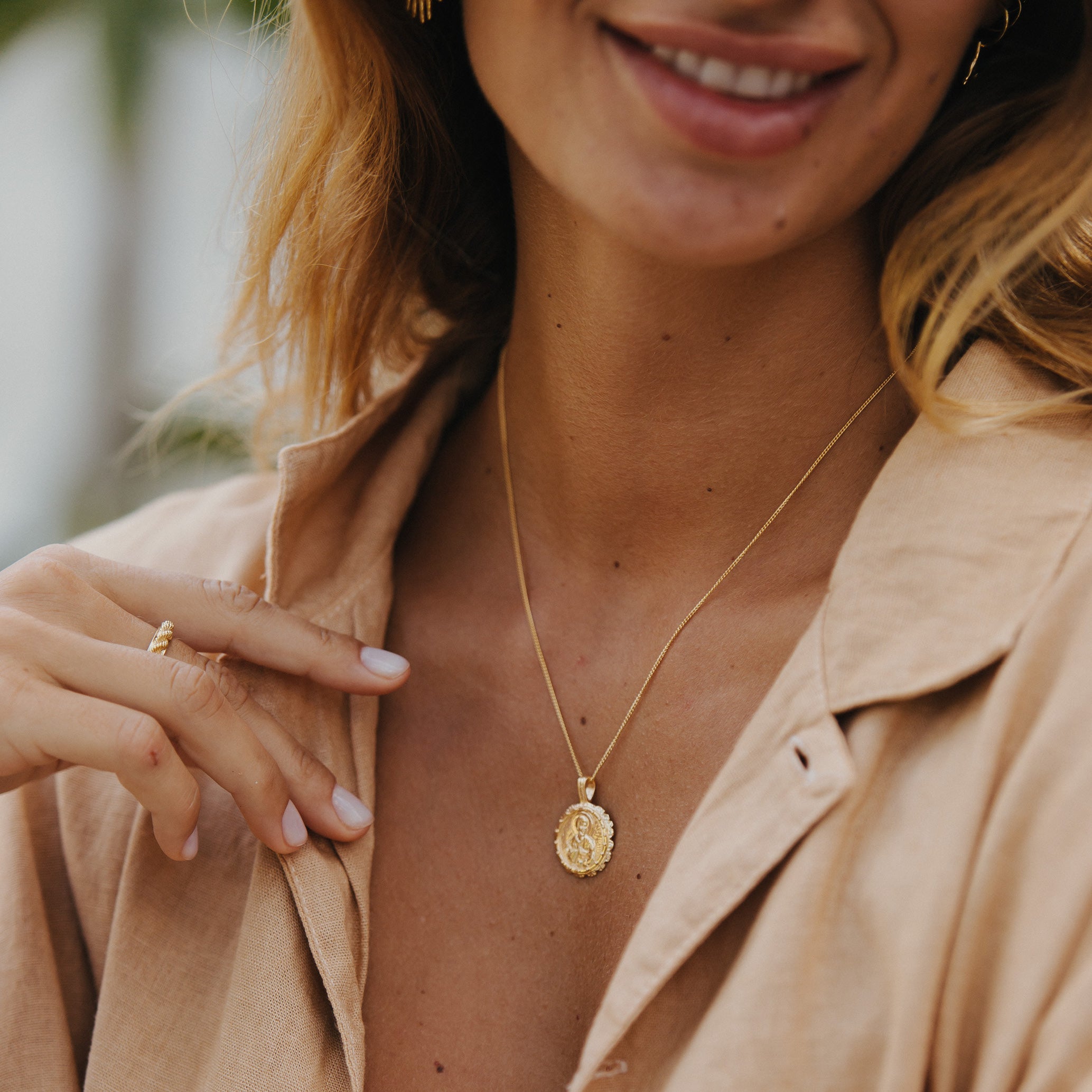 A woman wearing layered gold necklaces, including a saint pendant and a smaller medallion, paired with a gold ring and a beige blouse