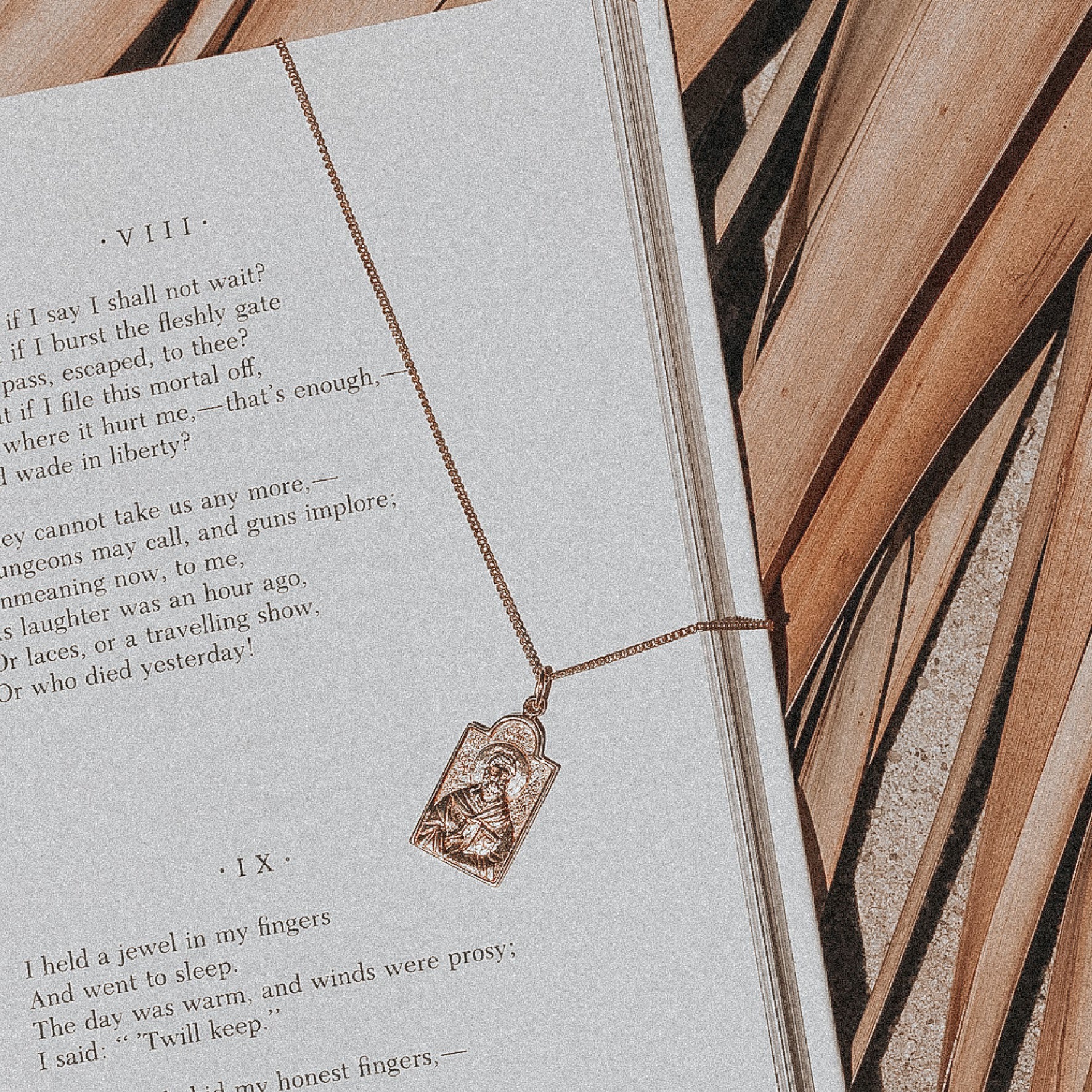 A gold necklace with a rectangular religious figure pendant, laid on an open book displaying poetry.