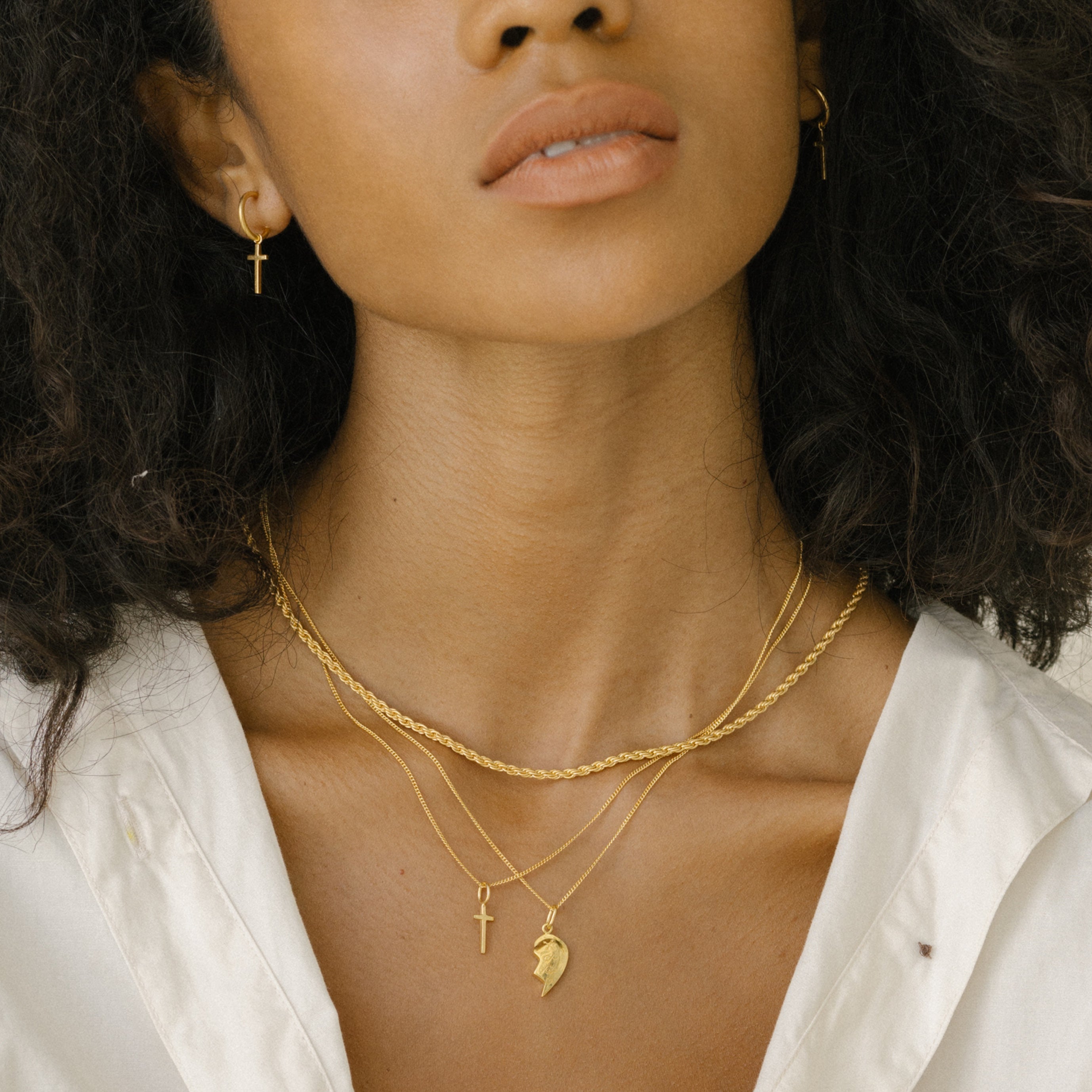 A woman wearing gold hoop earrings with small cross pendants and layered gold necklaces with cross and charm pendants, paired with a white shirt.