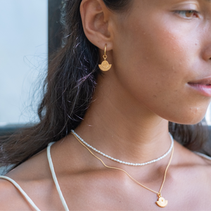  A close-up of a woman wearing gold sunburst earrings, a gold necklace with a sunburst pendant, and a pearl necklace.