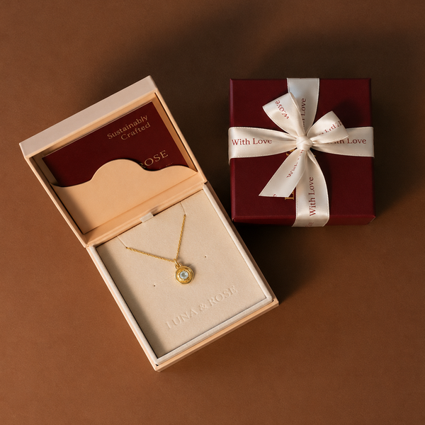 A gold necklace with a small round pendant displayed in an open cream-colored jewelry box, alongside a dark red gift box tied with a creme ribbon